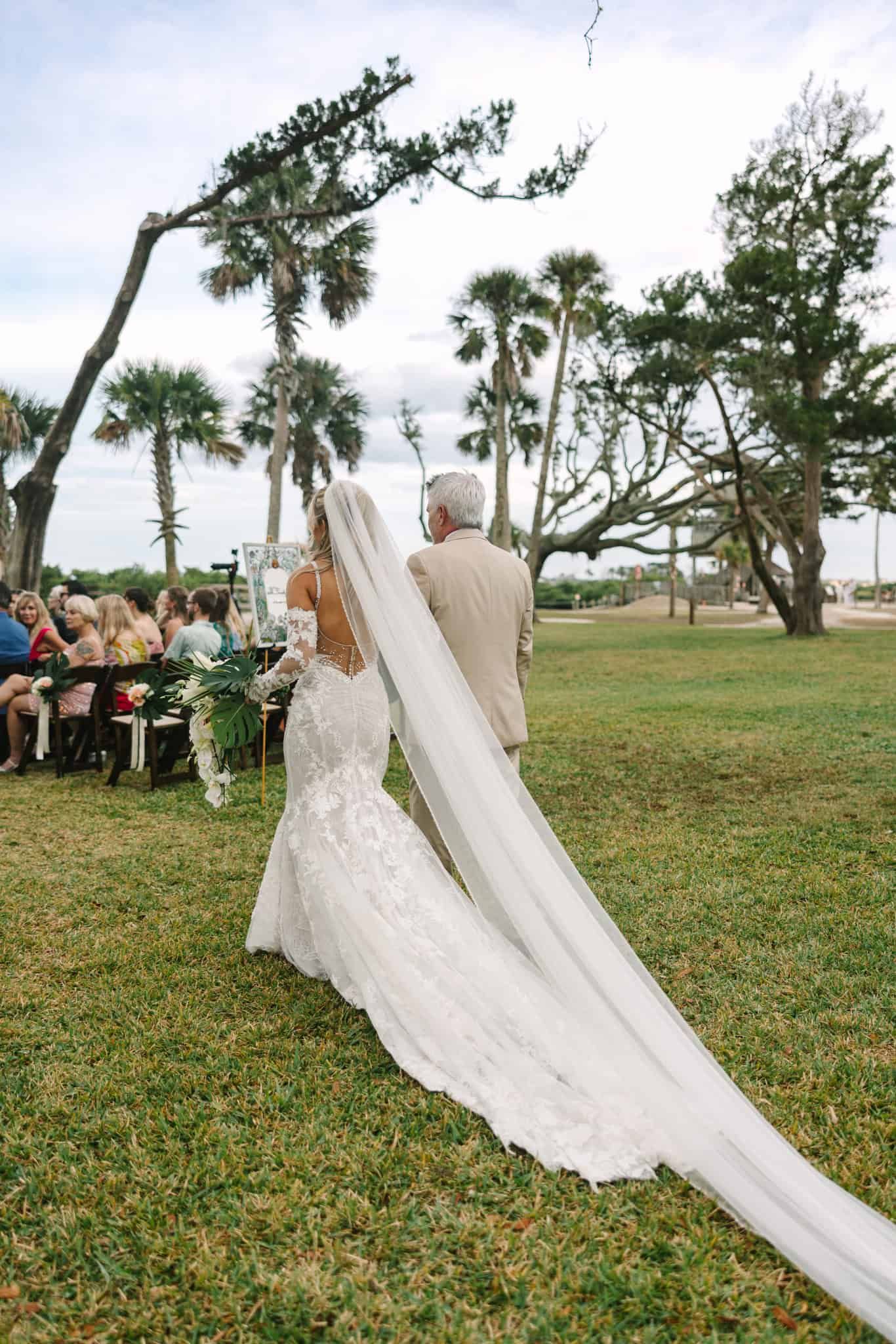 bride being walked down the aisle by her father from behind, veil and train flowing behind her