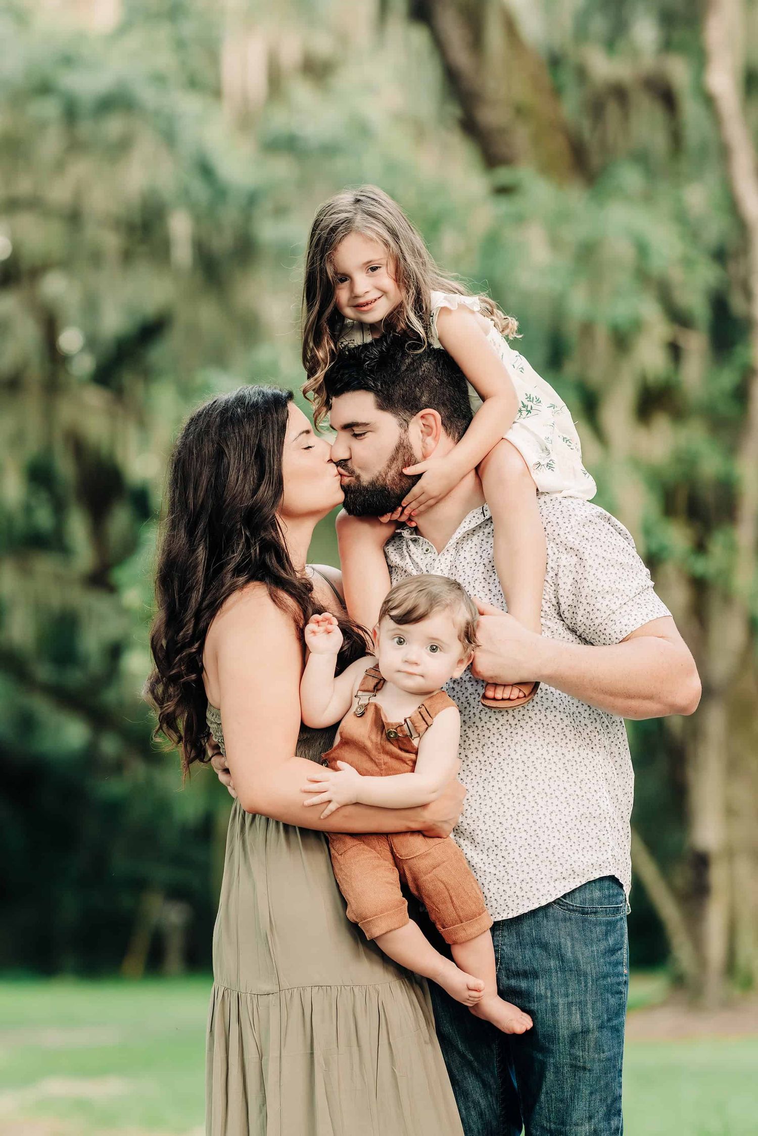 mom and dad give a kiss as mom holds toddler boy and daughter is smiling on dads shoulders
