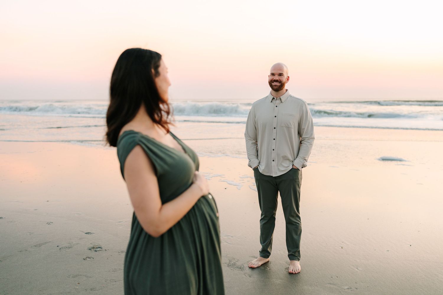 Pregnant woman in green dress and husband standing at Ponte Vedra beach sunset during maternity photo session, looking lovingly at wife