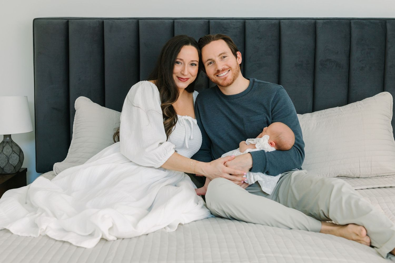 A happy couple sitting on a bed holding their newborn baby, smiling warmly in a bright, modern bedroom.