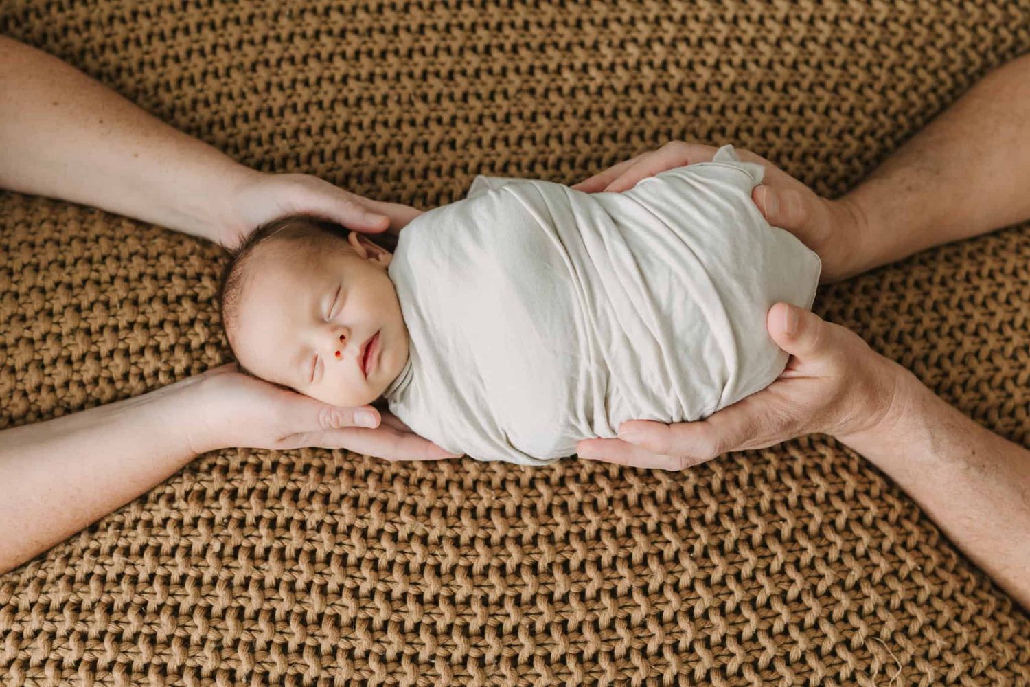 newborn baby swaddled up with parents hands holding him from above and below