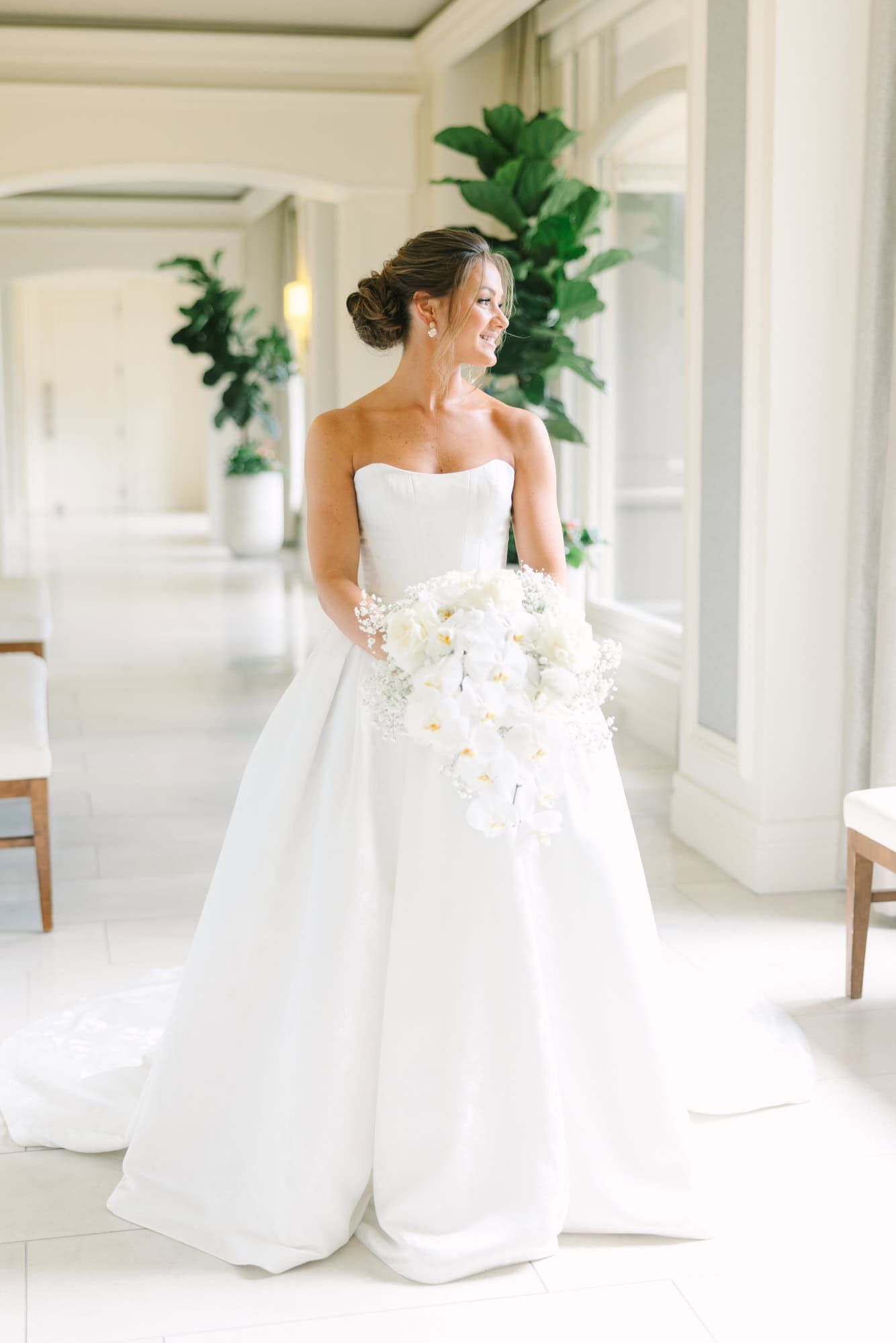 bride standing in a white hallway holding her bouquet and looking out the window to the right