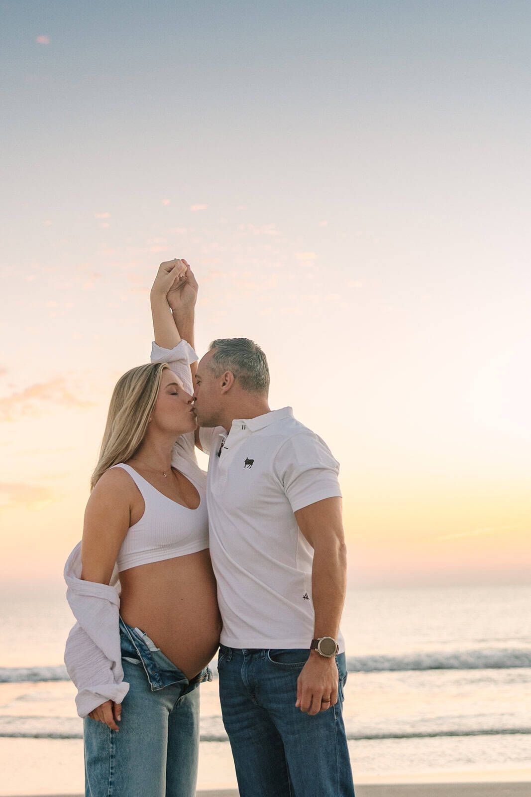 mom and dad kissing with moms pregnant belly out and hands overhead. pretty sunrise water and sky in the background