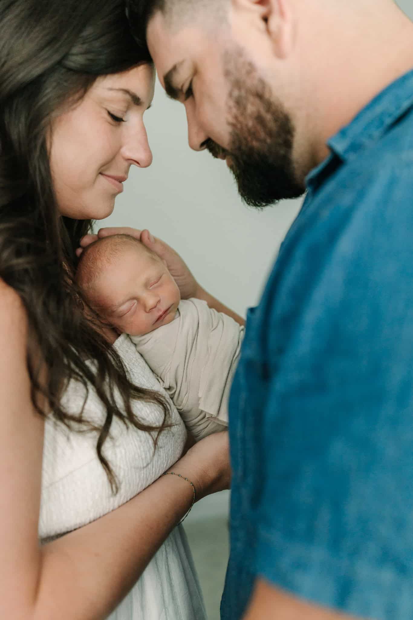 New parents tenderly touch foreheads while cradling their sleeping swaddled newborn baby together.