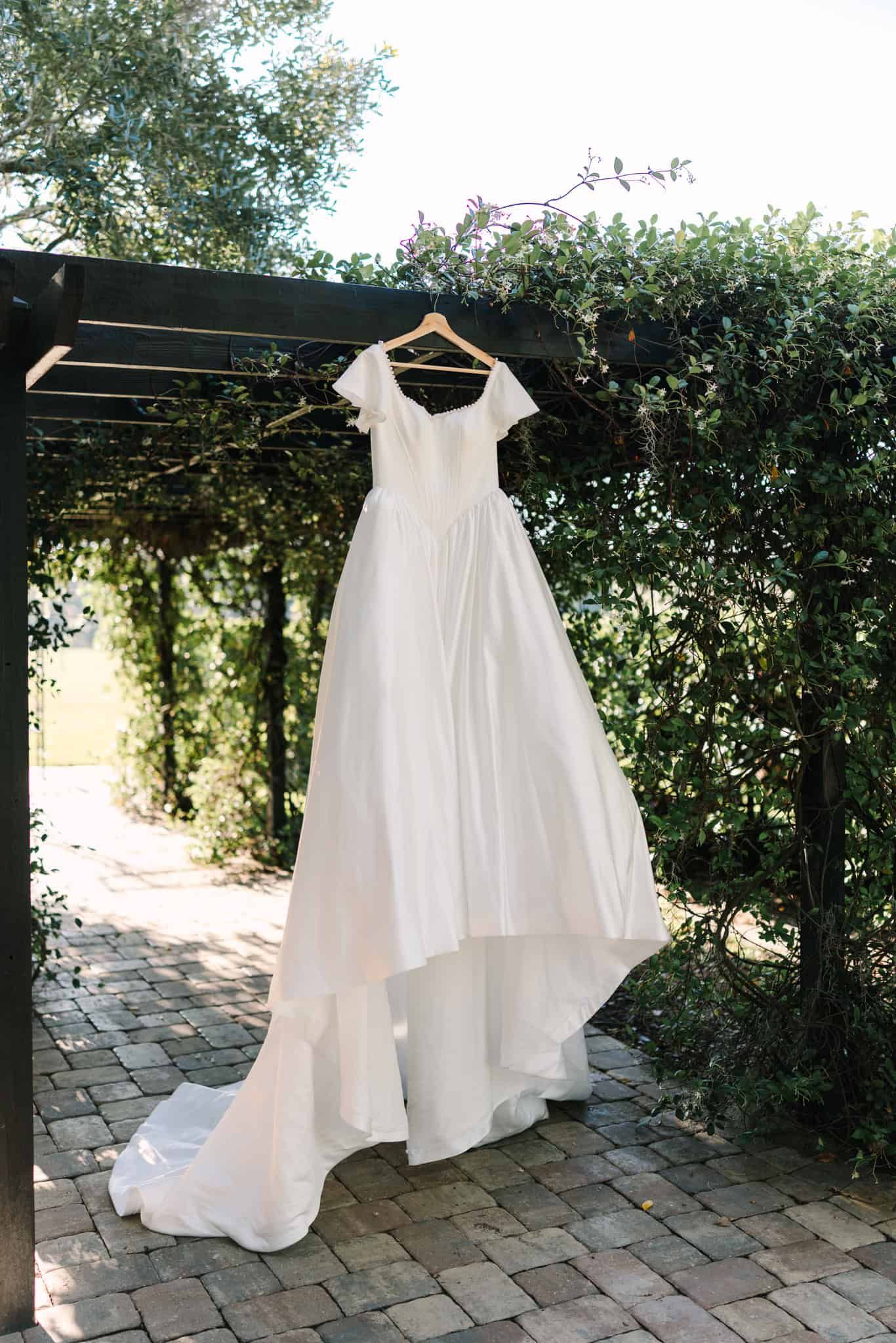 White A-line wedding dress with cap sleeves and train hanging on wooden hanger under garden pergola.