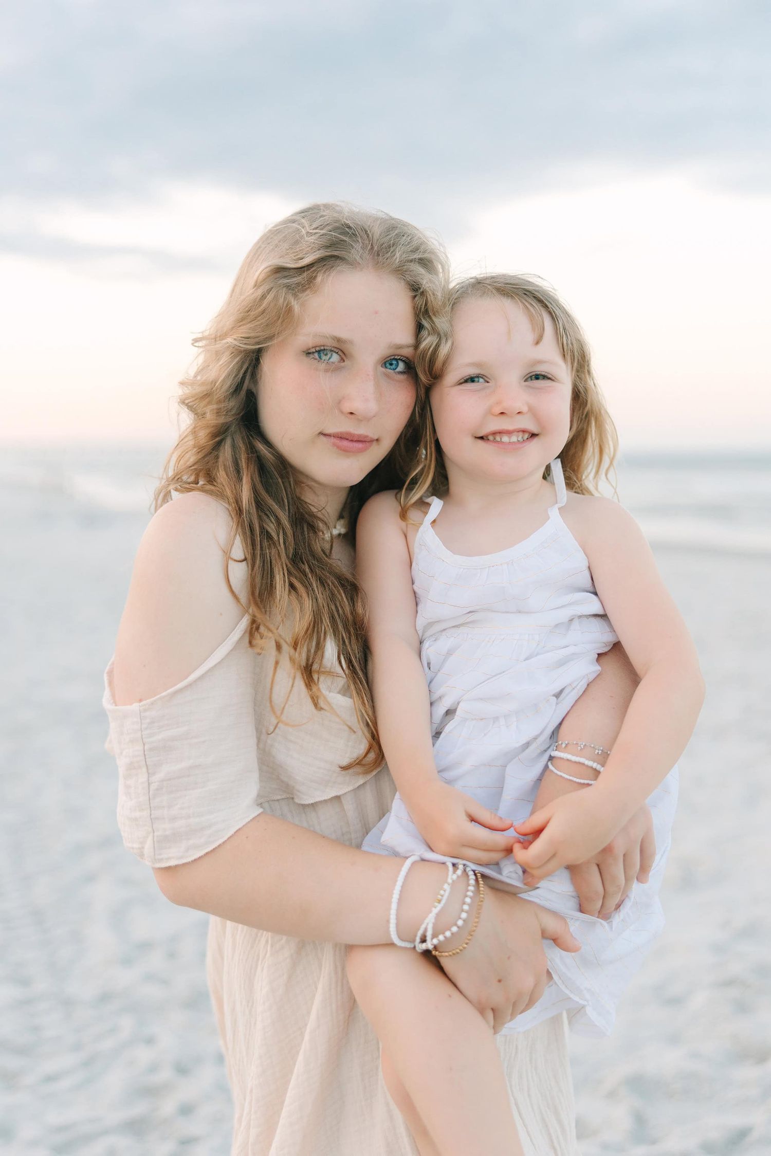 older daughter holding younger sister with bright blue and green eyes at the beach