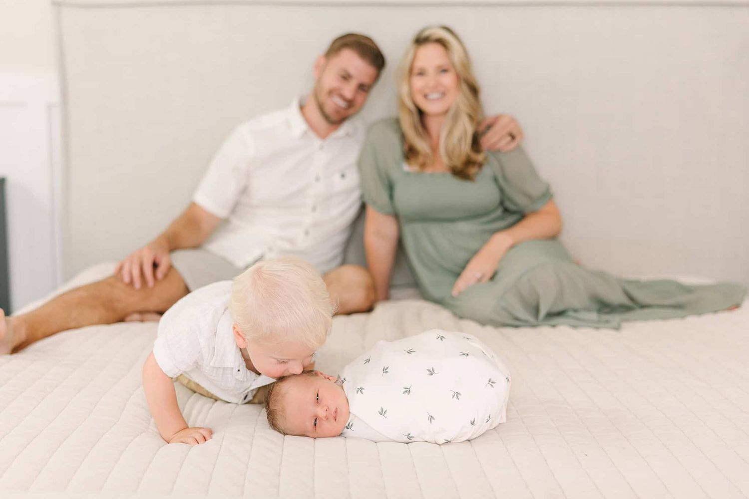 mom and dad sitting on their bed with toddler crawling up to the end of the bed to give newborn baby brother a kiss on his cheek