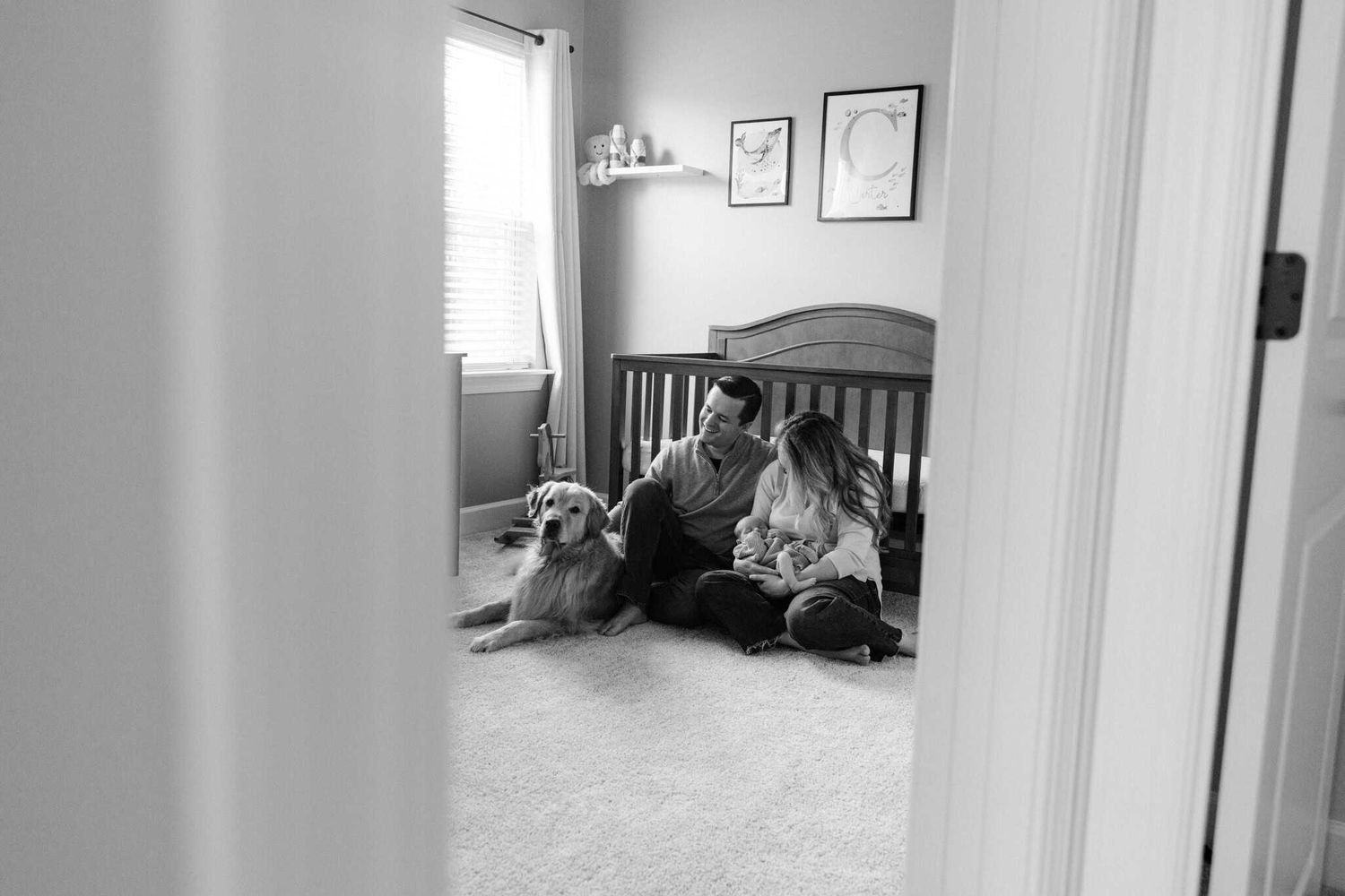 black and white photo peeking into the nursery of mom and dad holding newborn baby with dog also in the nursery