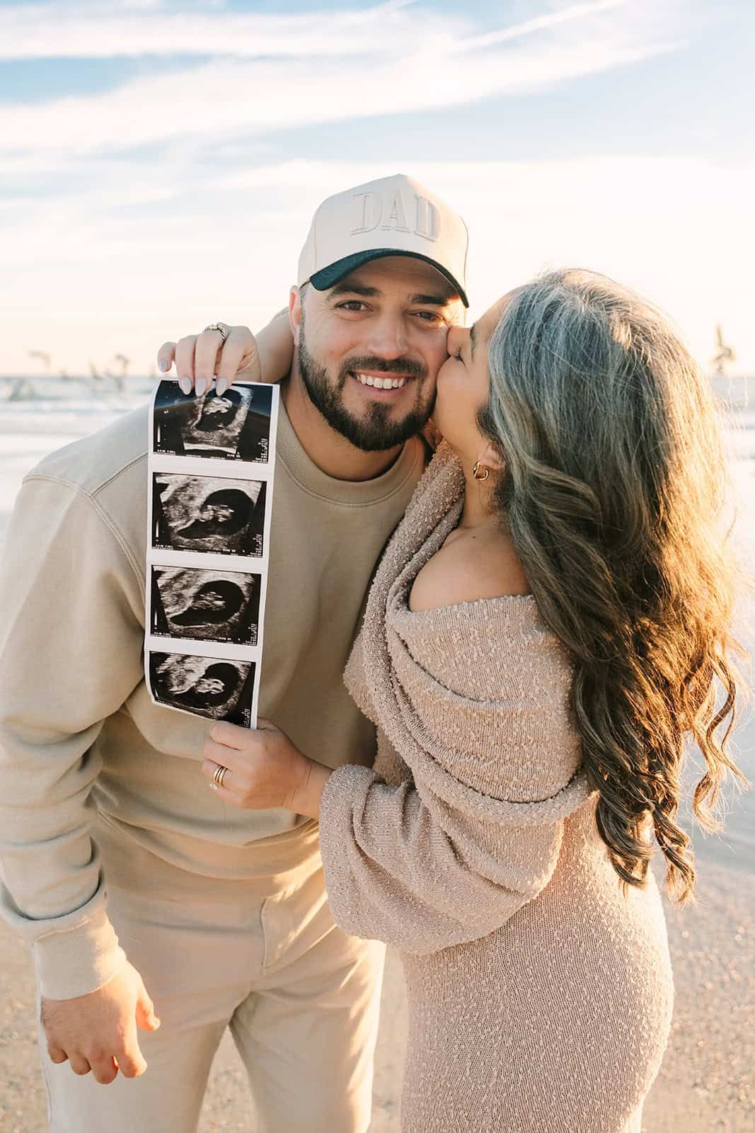 new mom giving dad a kiss on the cheek, holding the ultrasound, and he has on a dad baseball hat