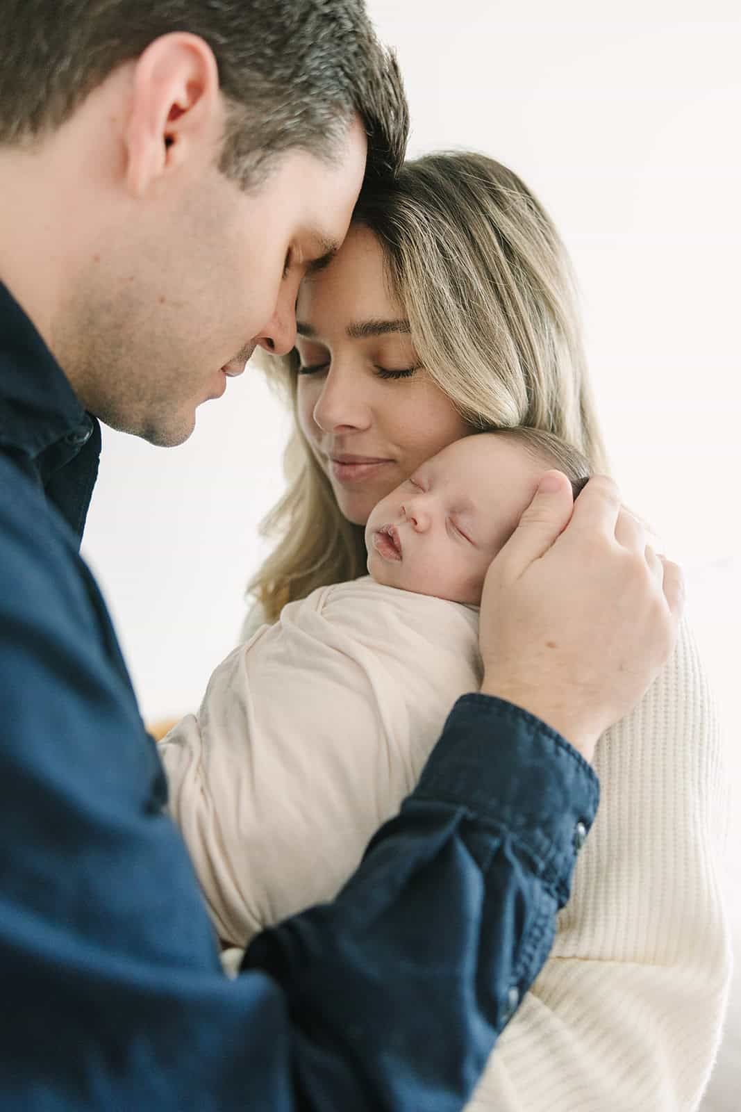 mom and dad snuggling newborn baby in Jacksonville fl with natural light hitting their faces from a big window