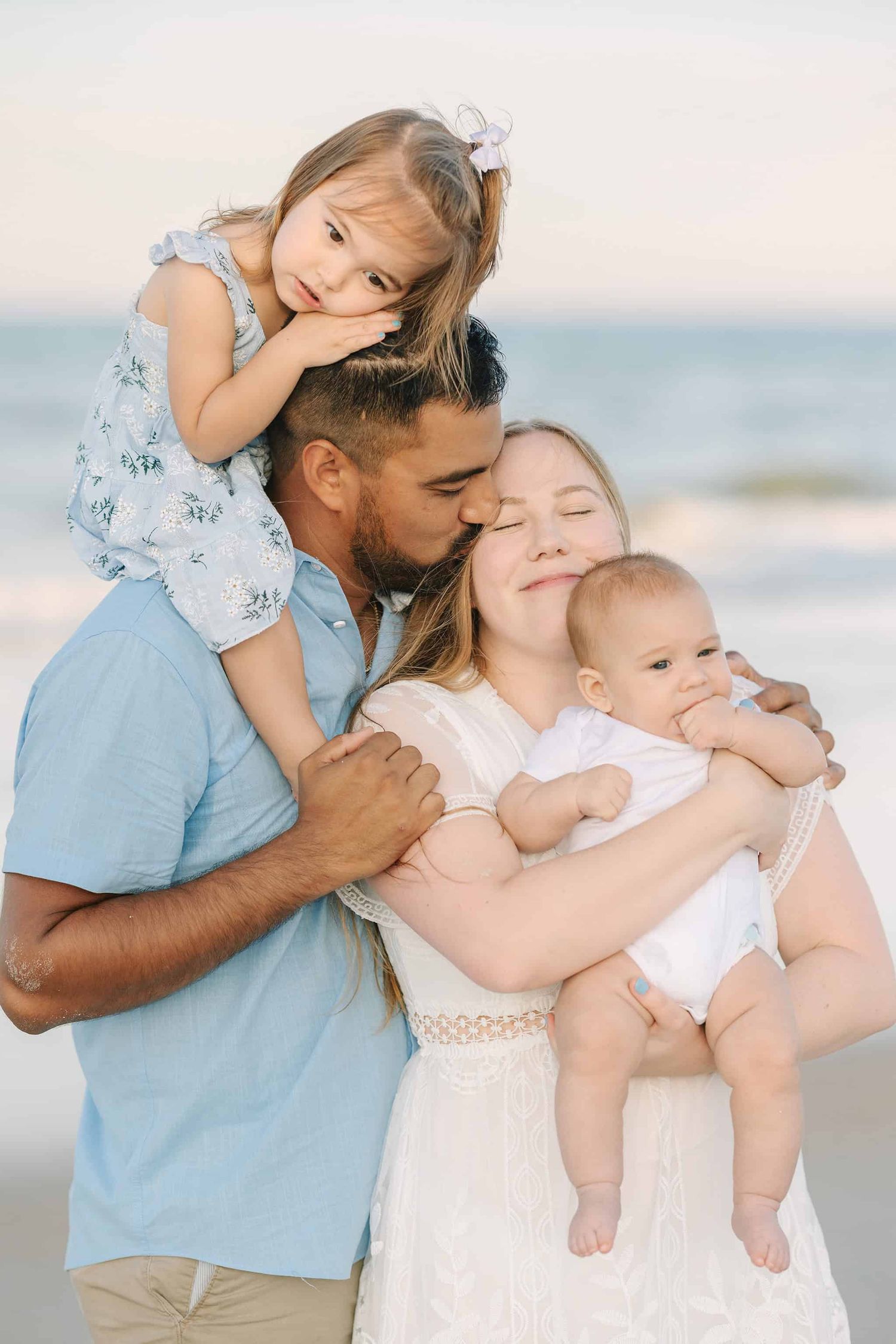dad kissing mom on the head while she snuggles with baby boy and toddler girl is on dads shoulders. all wearing blue and white