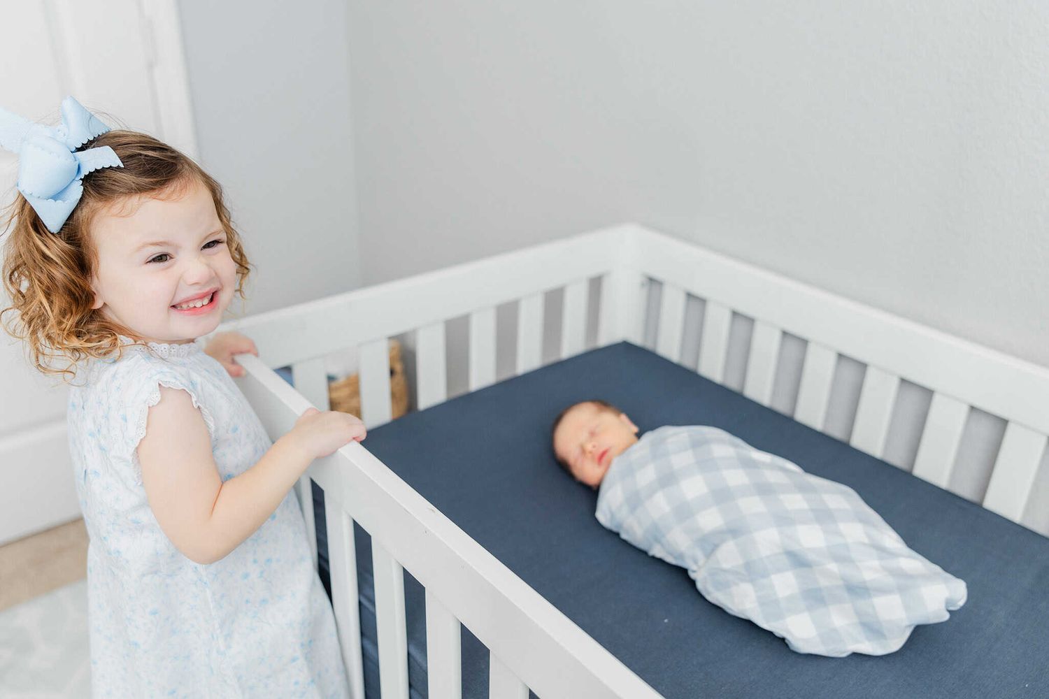 big sister climbing on crib smiling at camera with newborn baby brother in the crib