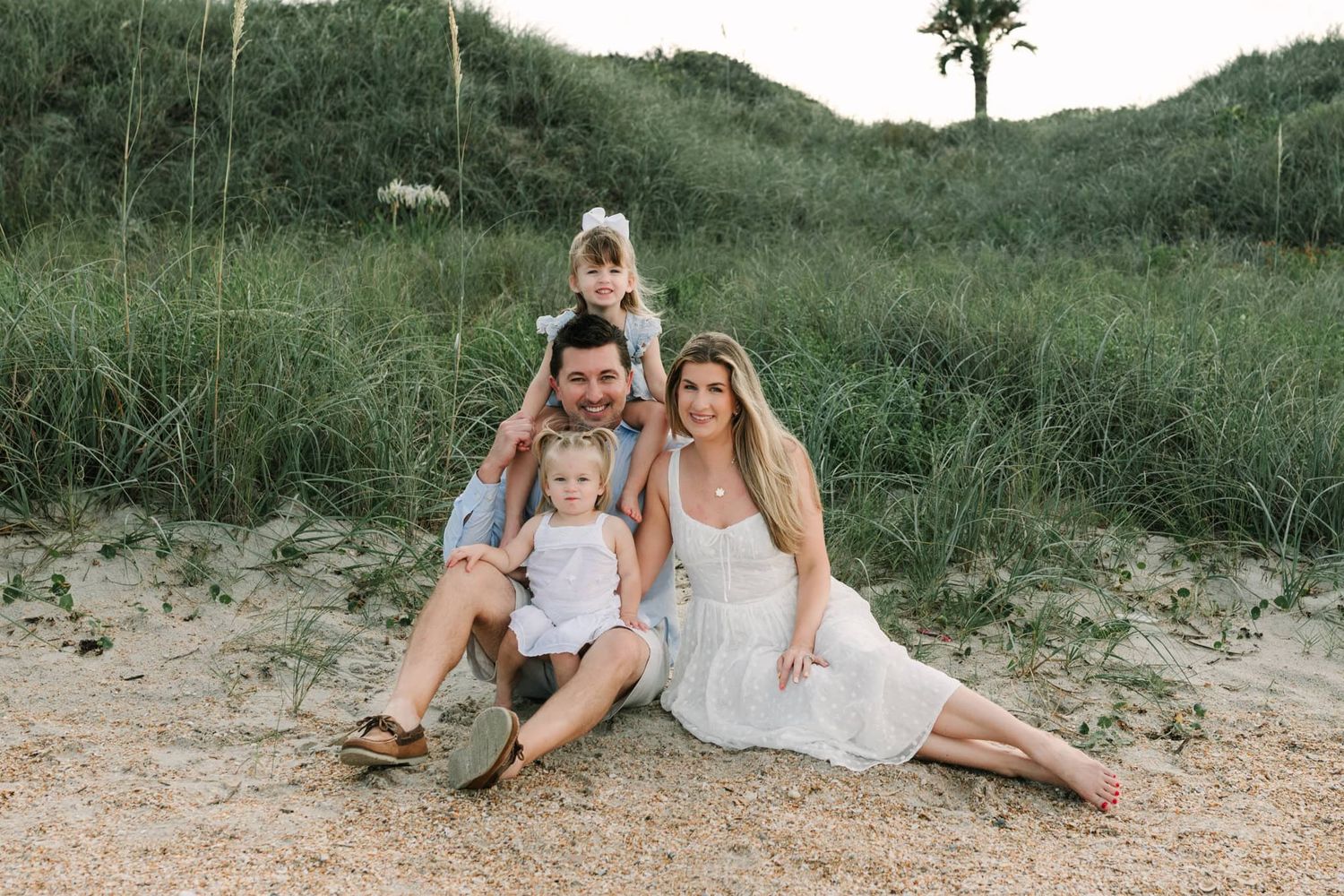 Happy family of four posing on sandy jacksonville beach, fl dunes with lush green grass, parents smiling with two young children.