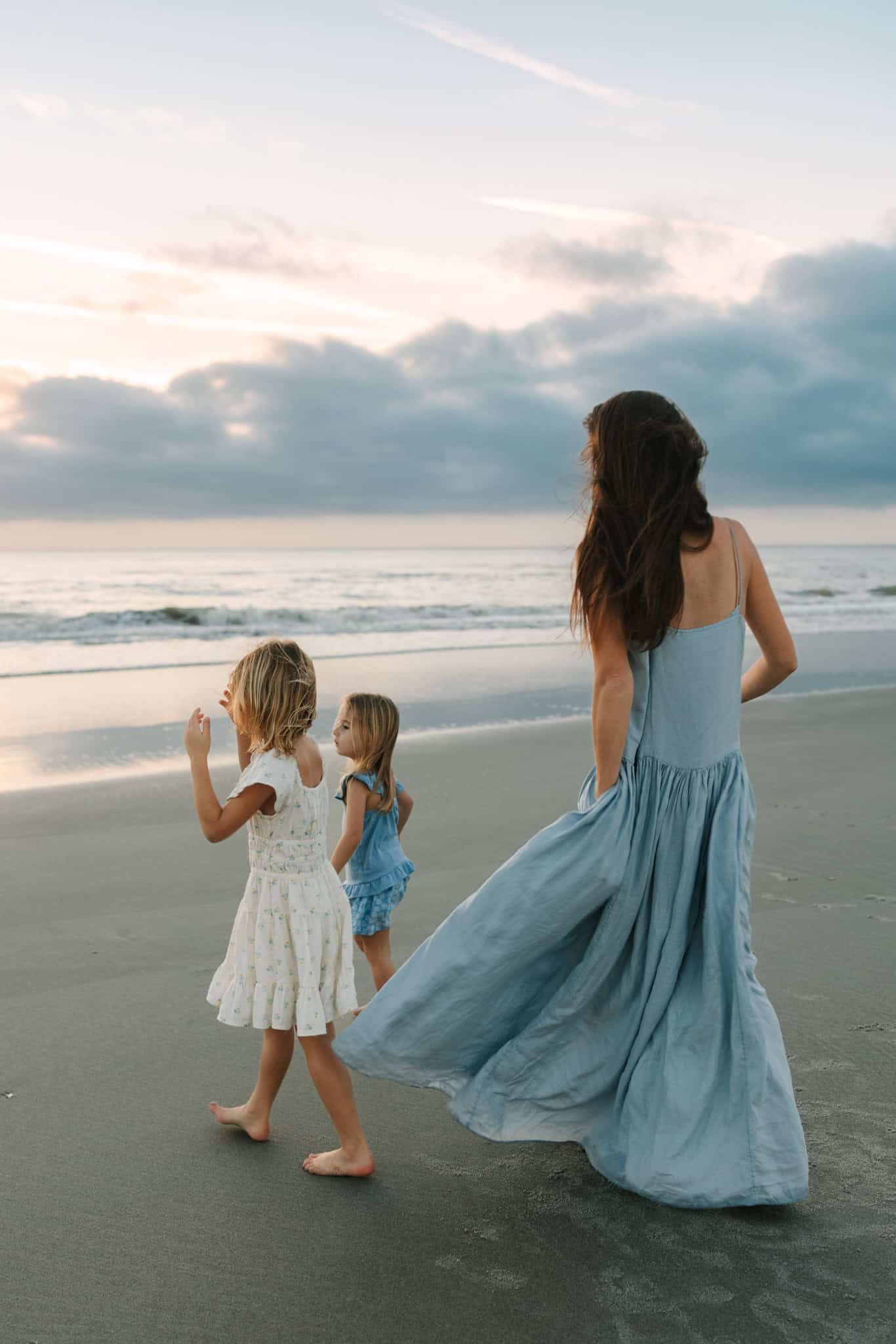 mom walking down Jacksonville beach towards the ocean with her two daughters and dresses blowing in the wind