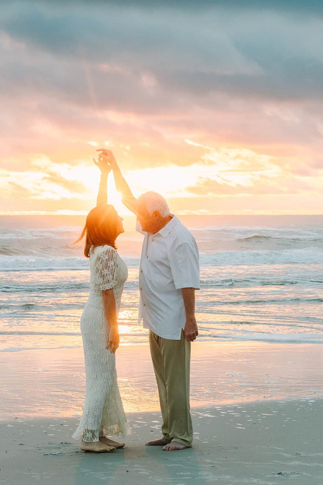 Elderly couple dancing on beach at sunset, woman in white lace dress, man in white shirt, waves and colorful sky behind them.