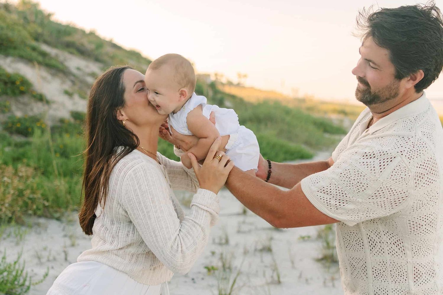 dad air planing baby girl to give mom a big kiss with sunset and dunes in the background