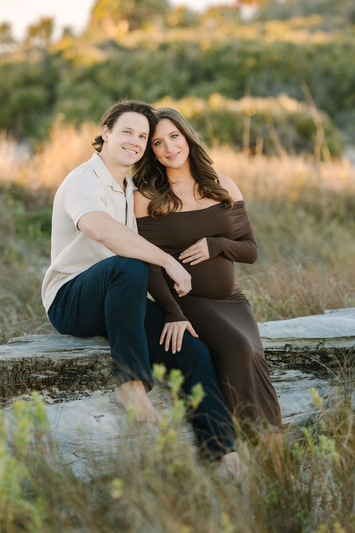 A couple sits on a log in Ponte Vedra beach dunes, the woman in a brown off-shoulder maternity dress, both smiling warmly