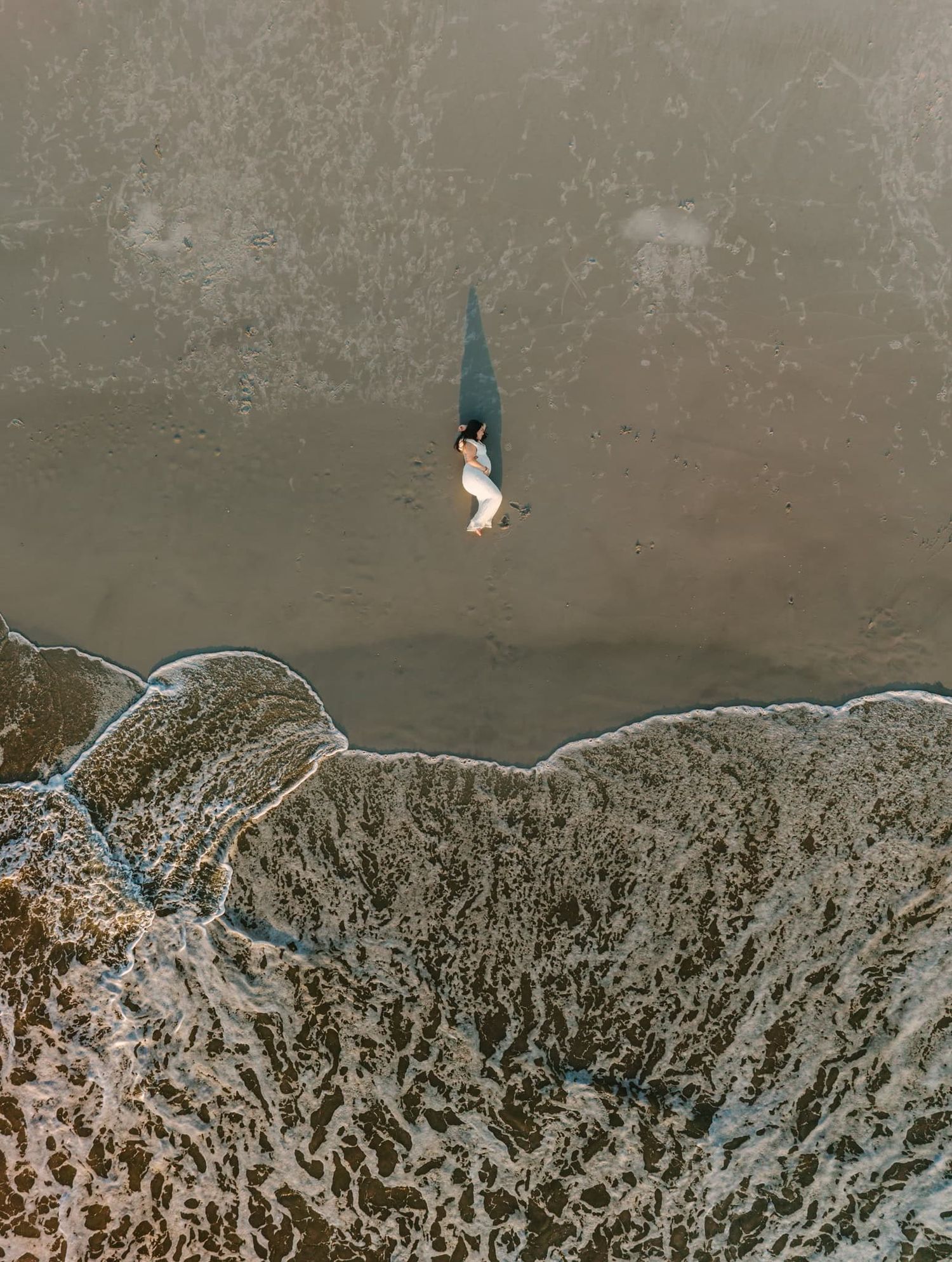Aerial drone view of a pregnant mama in white dress laying on a sandy beach near ocean waves with long shadow.