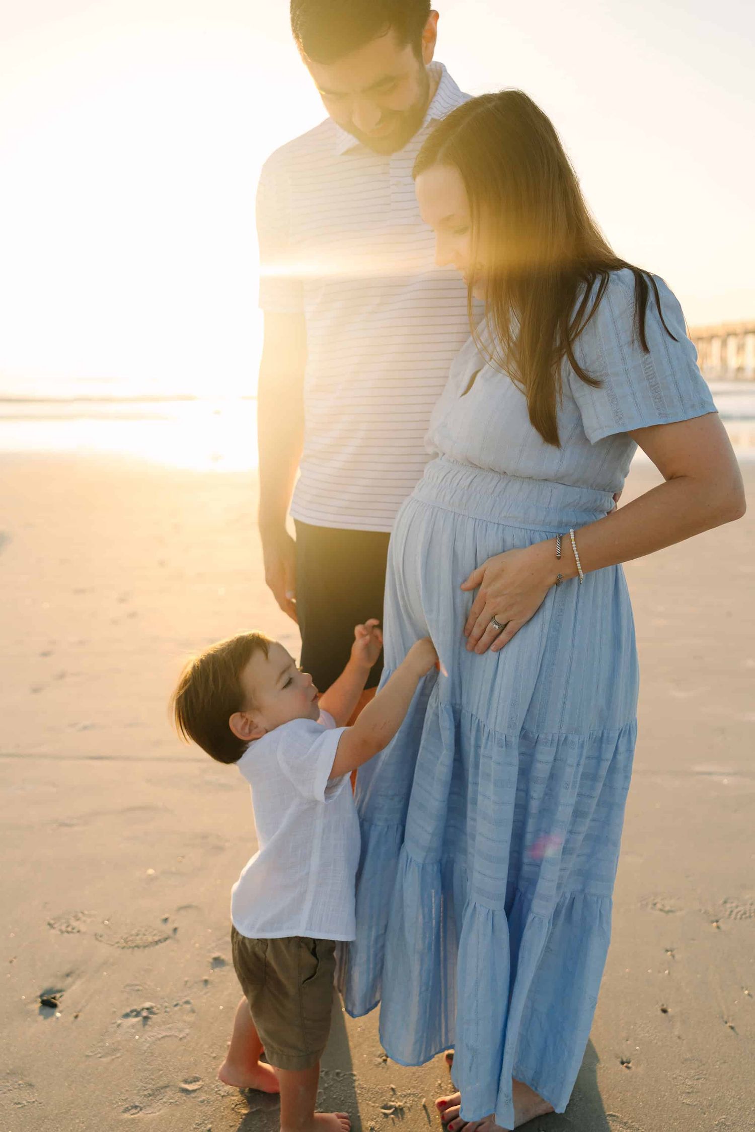 toddler son touching mom's pregnant belly with sunrise peeking out from behind them at the beach