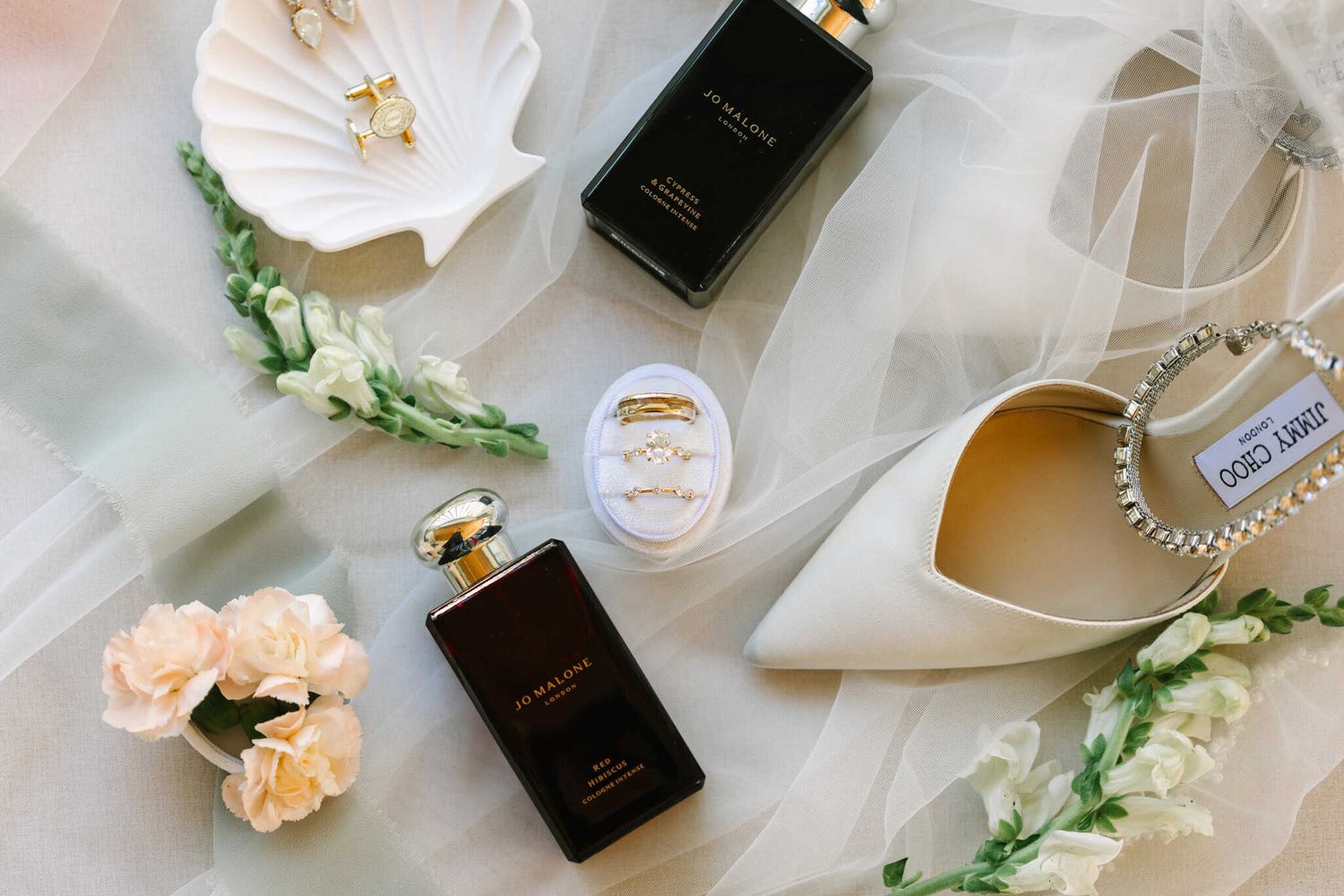 Bridal flat lay with Jimmy Choo heels, perfume bottles, pearl ring, and pink flowers on white veil.
