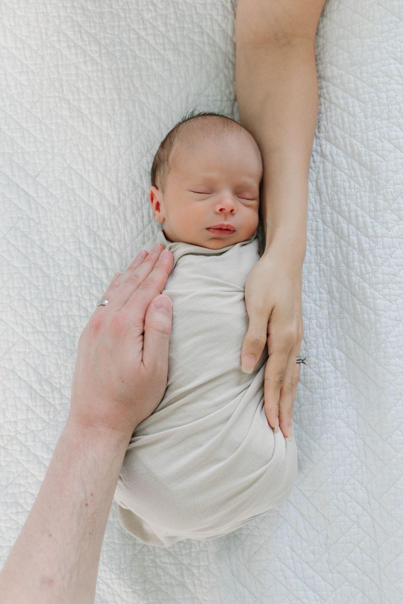 Newborn baby swaddled in cream wrap sleeping peacefully while mom and dad's hands gently cradle him on white quilted blanket.