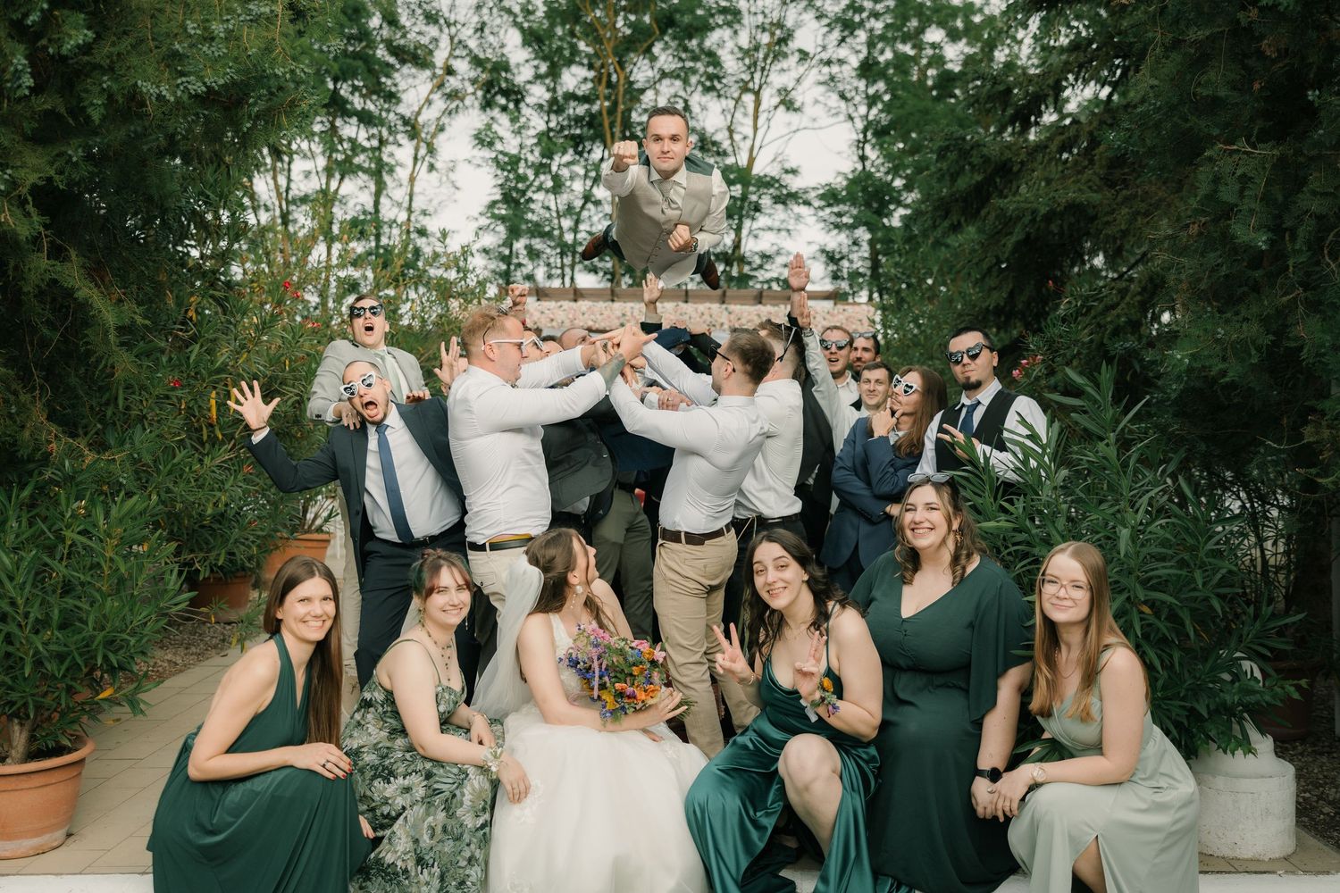 Wedding party in green dresses and khaki suits celebrating outdoors, with groom tossed in air among lush greenery.