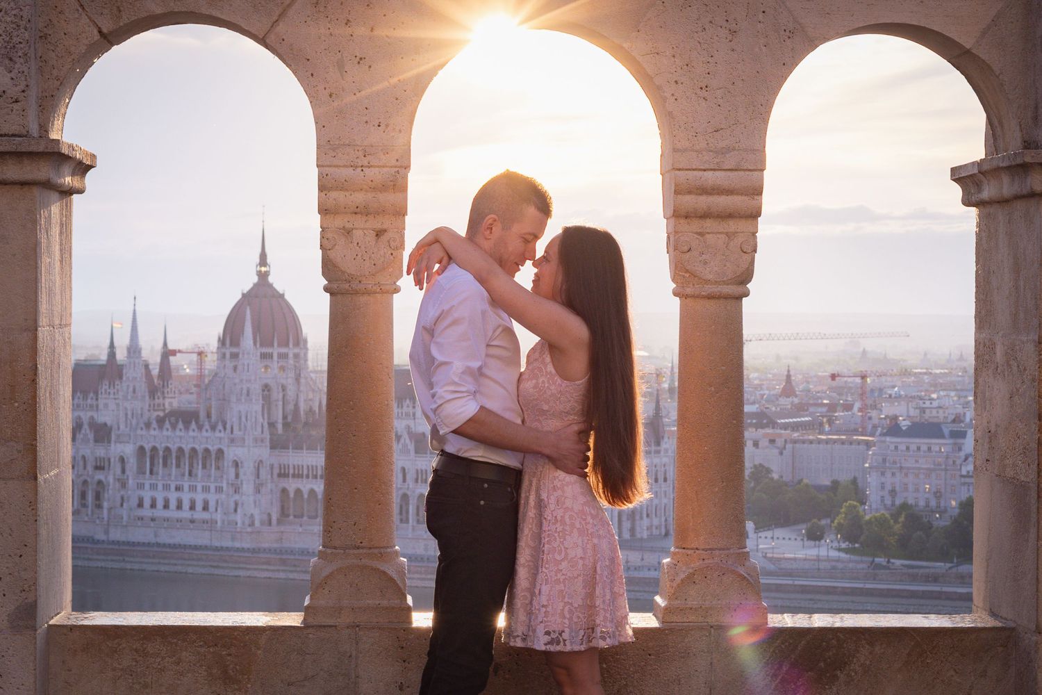 Couple silhouetted against arched windows overlooking Budapest Parliament building at sunset.