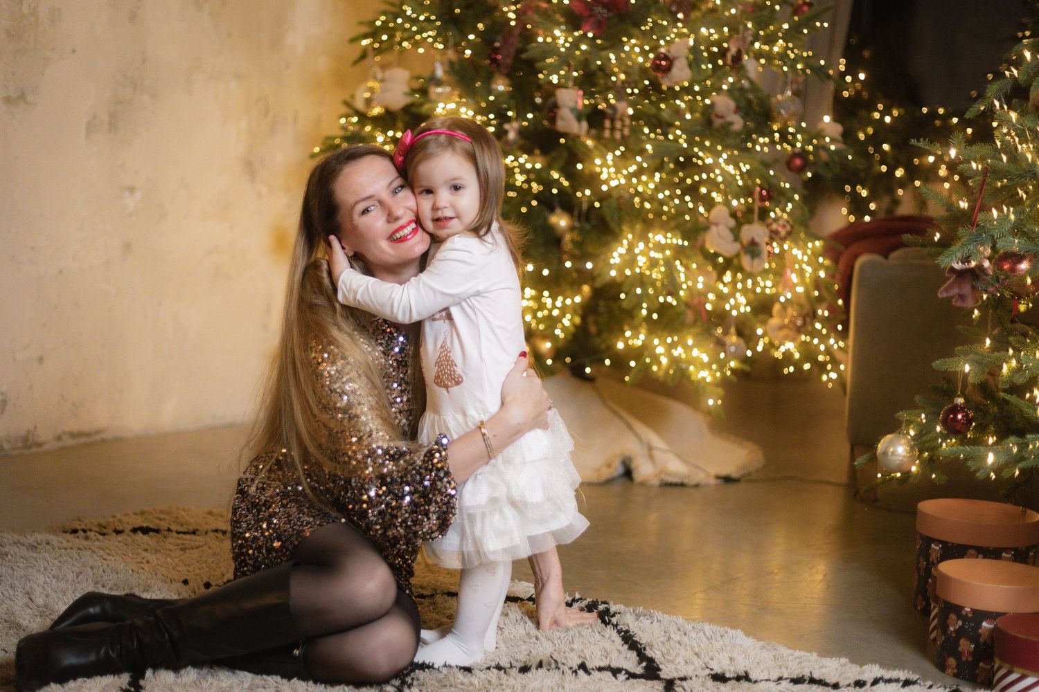 Woman in sequin dress hugs smiling toddler girl in white dress beside a glowing decorated Christmas tree.