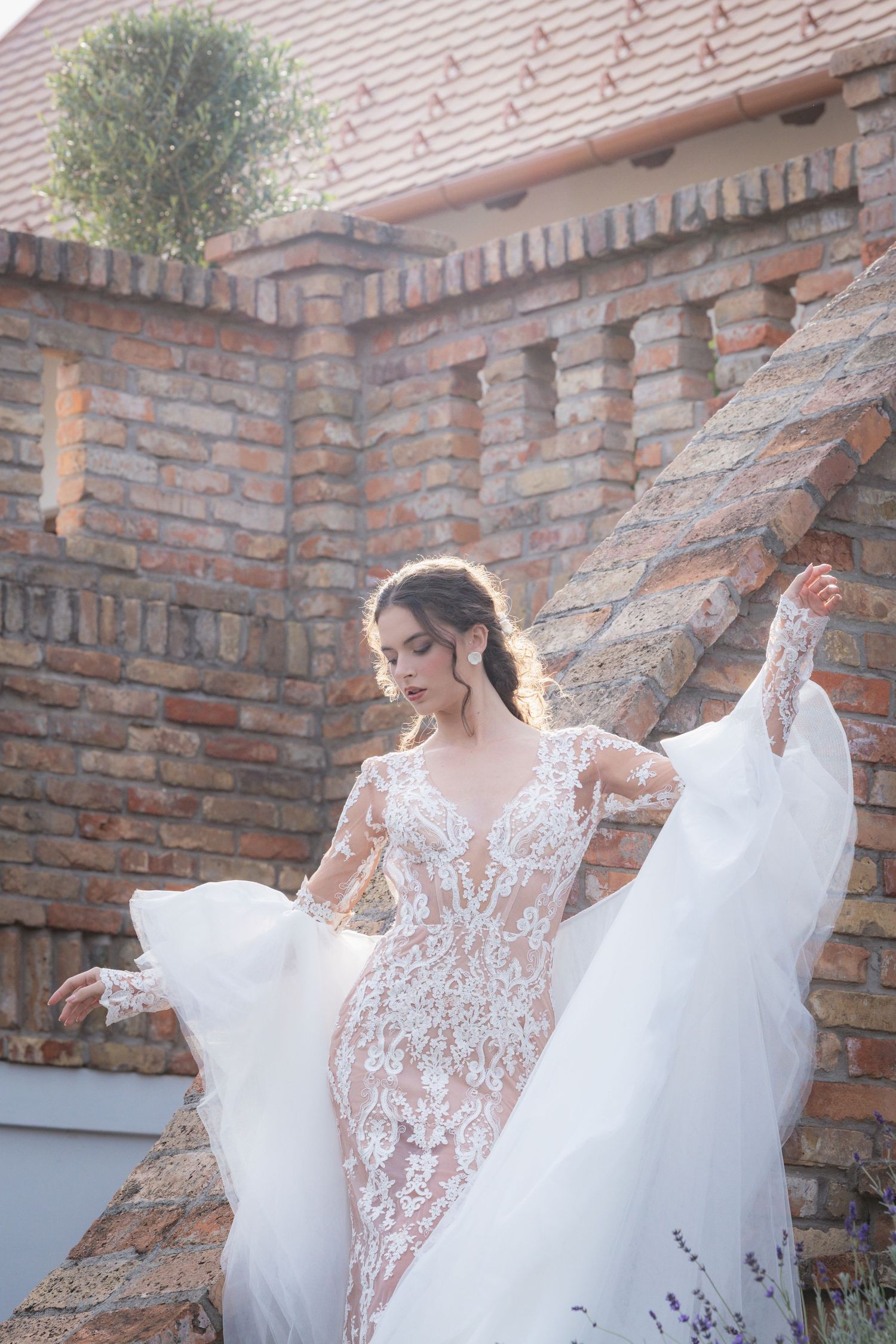 A bride in a flowing lace gown twirls gracefully against a rustic brick wall backdrop.