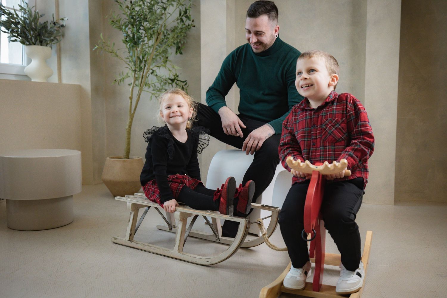Father watches two children in matching plaid outfits playing on wooden sled and rocking horse in bright modern room.