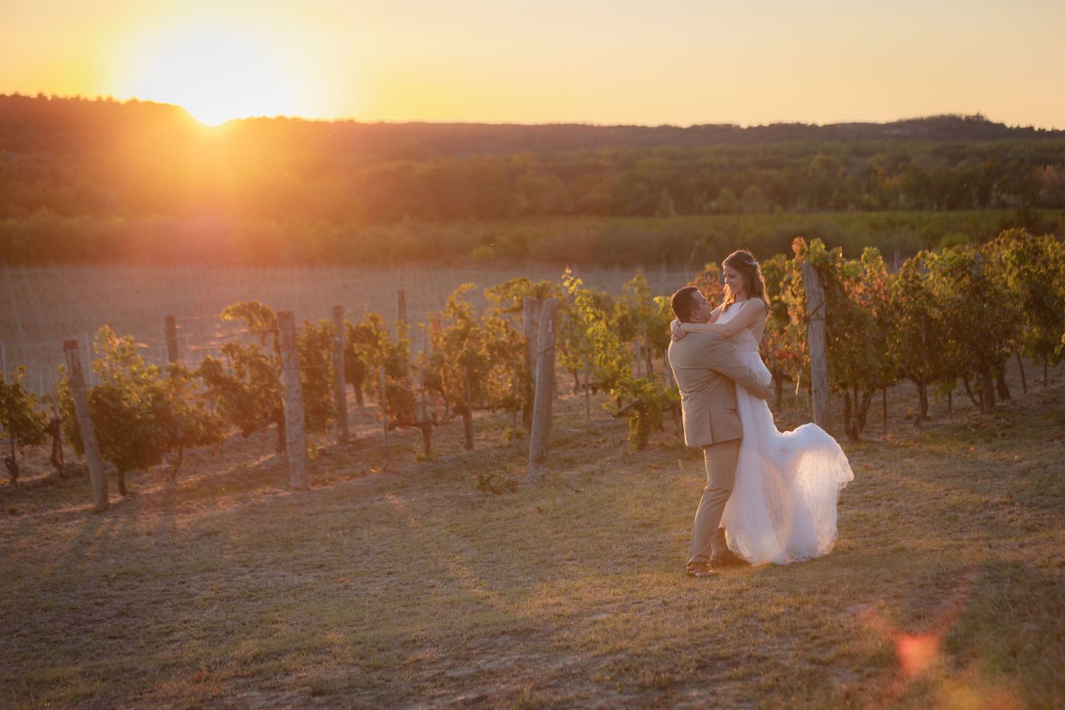 A couple walks through a vineyard at sunset with golden light streaming through the rows of grapevines.