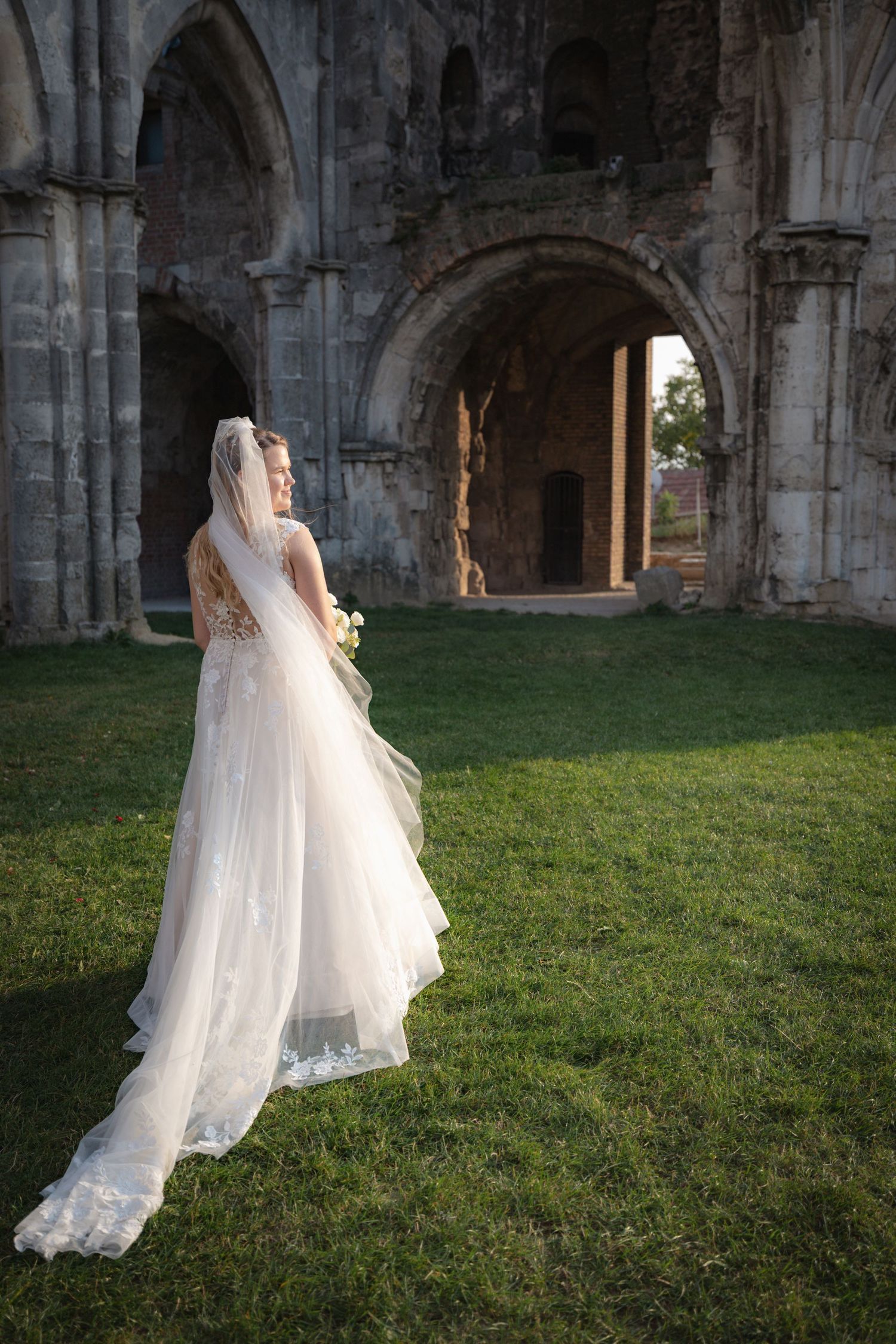 Flowing white wedding dress trails across green grass inside gothic stone church ruins.
