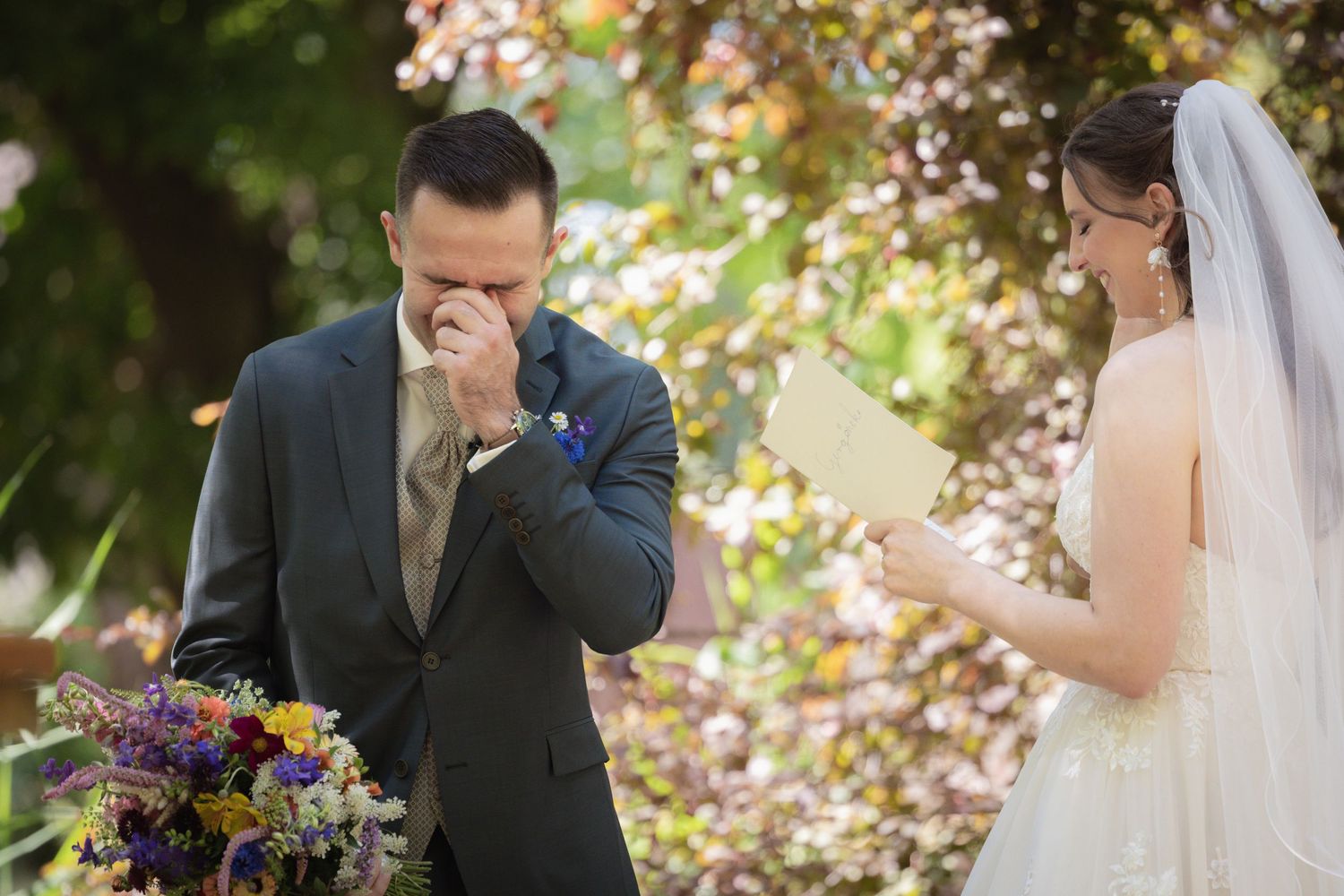 Emotional groom wiping tears as bride reads wedding vows outdoors, surrounded by colorful flowers and autumn foliage.