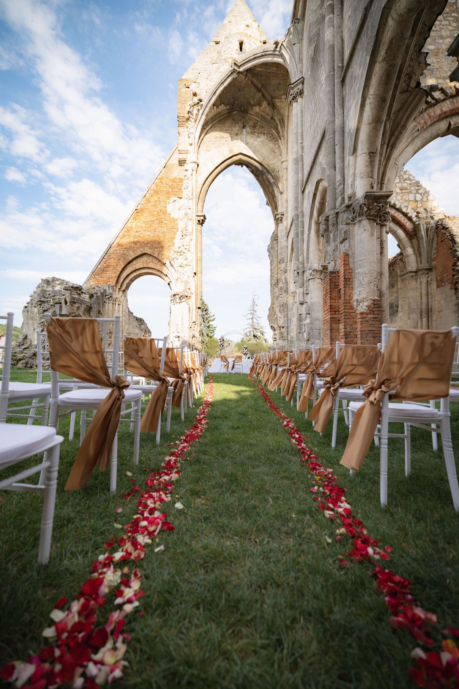 White chairs line a grassy aisle with red rose petals in ancient stone church ruins under blue sky.