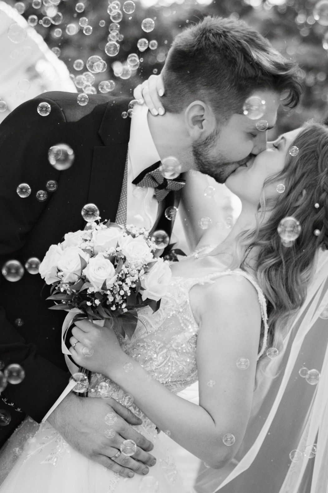 Black and white photo of a bride and groom kissing surrounded by bubbles, bride holding a white rose bouquet.