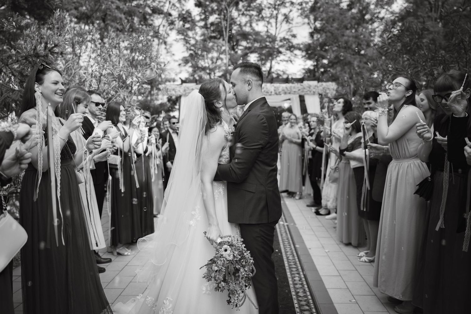 Black and white photo of newlyweds sharing a kiss while guests celebrate along the aisle at an outdoor wedding ceremony.