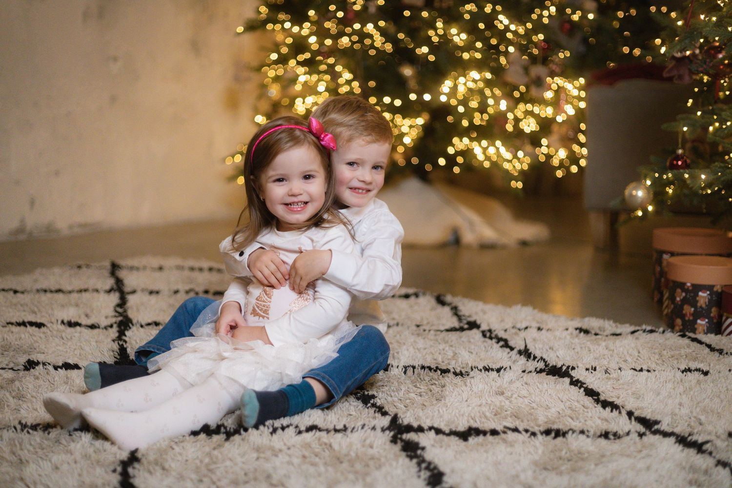 Two young children sitting on a rug hugging in front of a decorated Christmas tree with warm glowing lights.