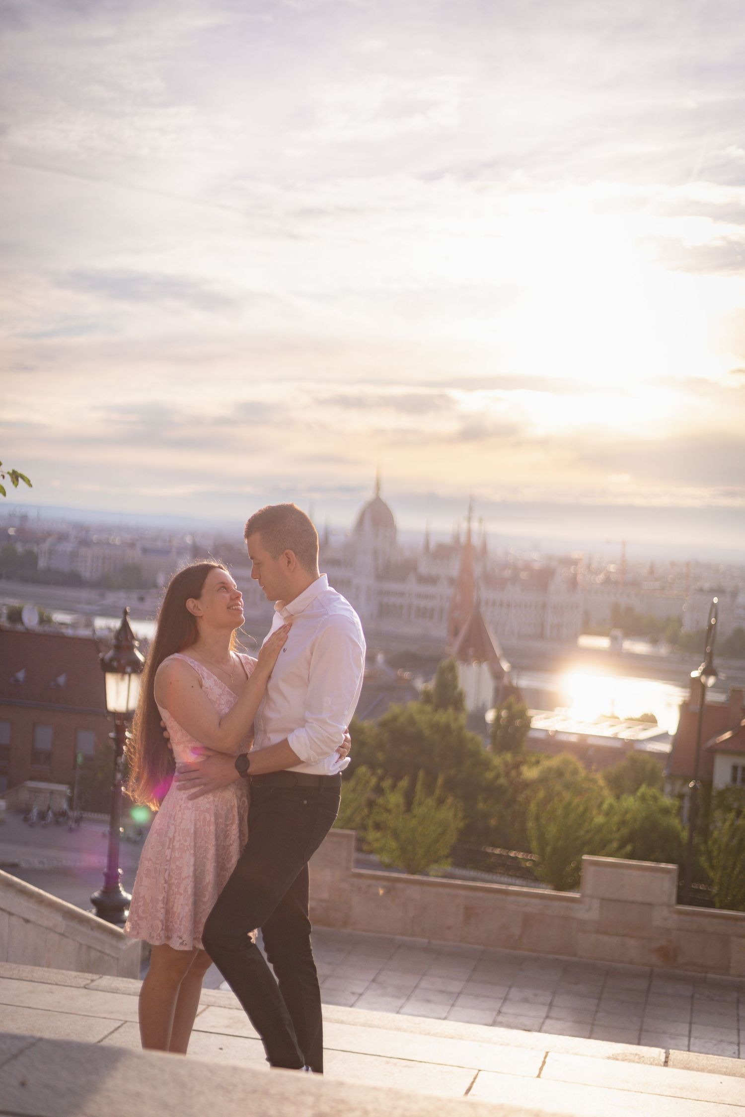 A romantic couple embraces during sunset overlooking Budapest's cityscape with the Parliament building visible in the distance.