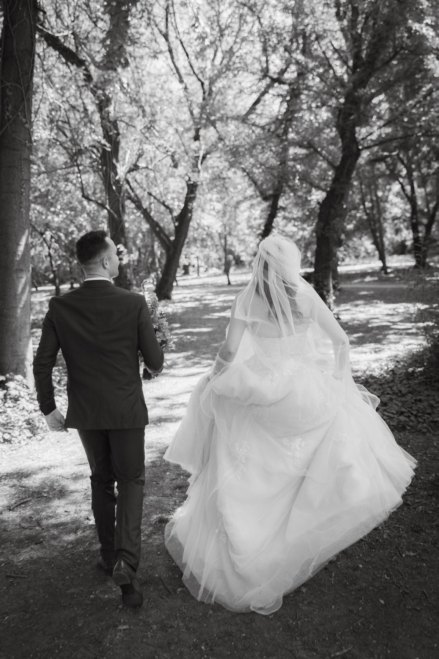 Black and white photo of a bride and groom walking hand in hand through a tree-lined park path on their wedding day.