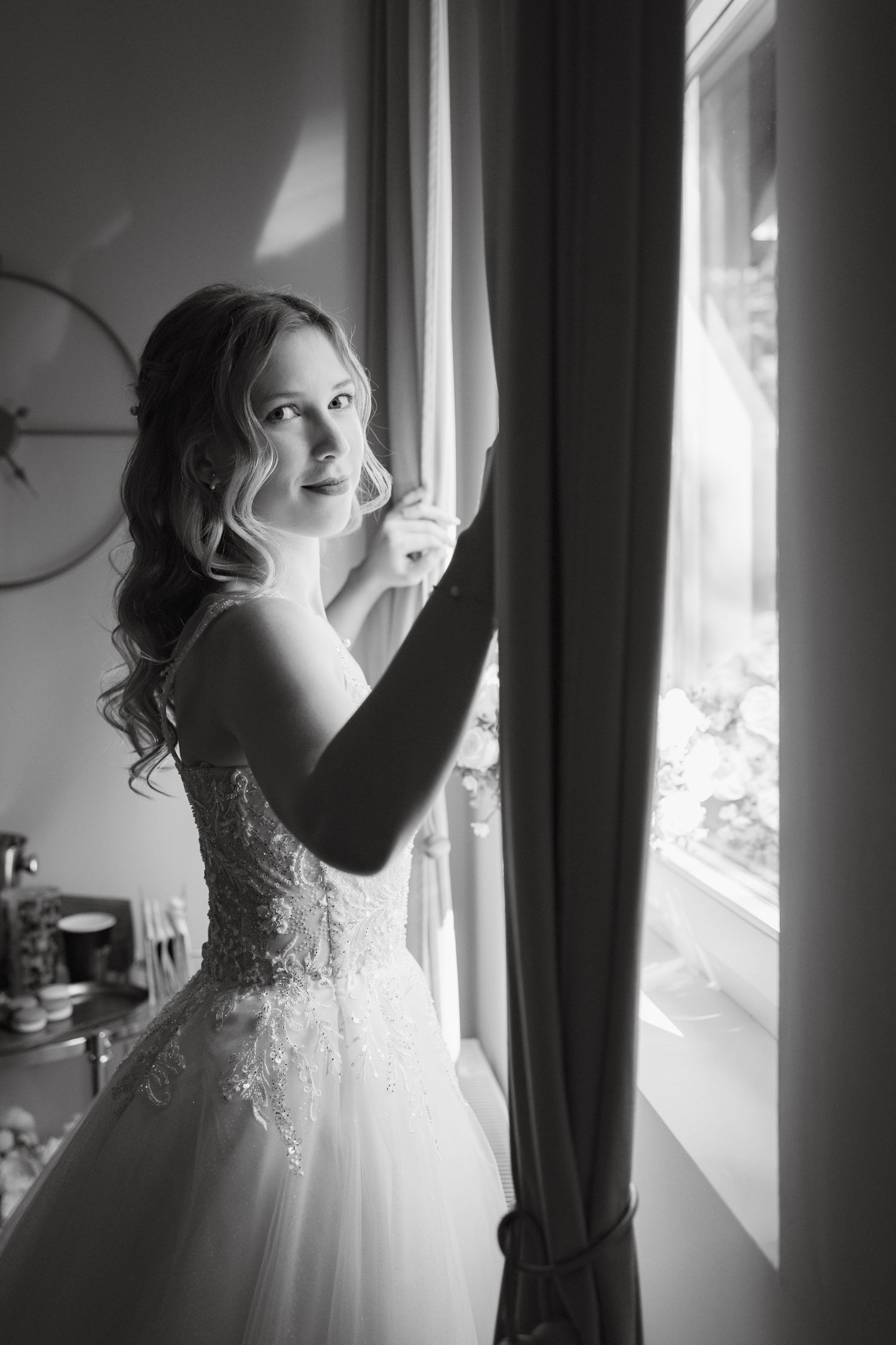Black and white photo of a bride in a lace wedding gown standing by a sunlit window holding a curtain.