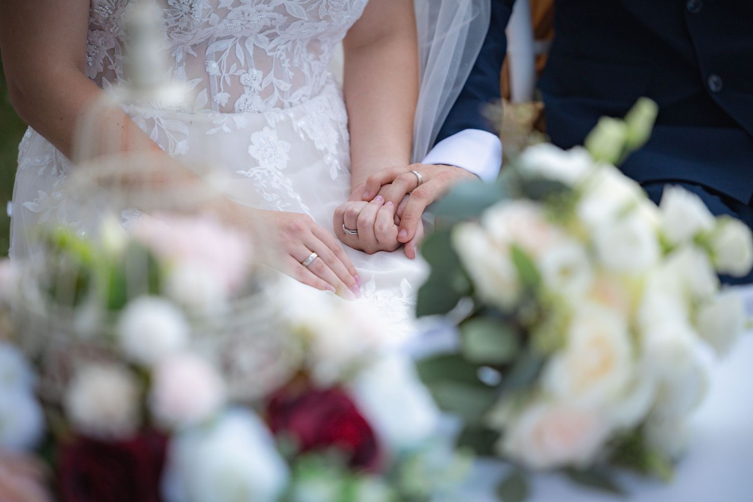 Close-up of hands with wedding rings among elegant white and red floral arrangements.