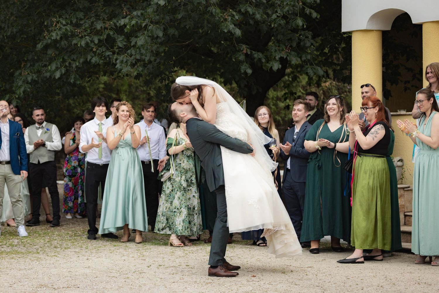 A groom dips his bride for a kiss as wedding guests in mint green dresses applaud outdoors.