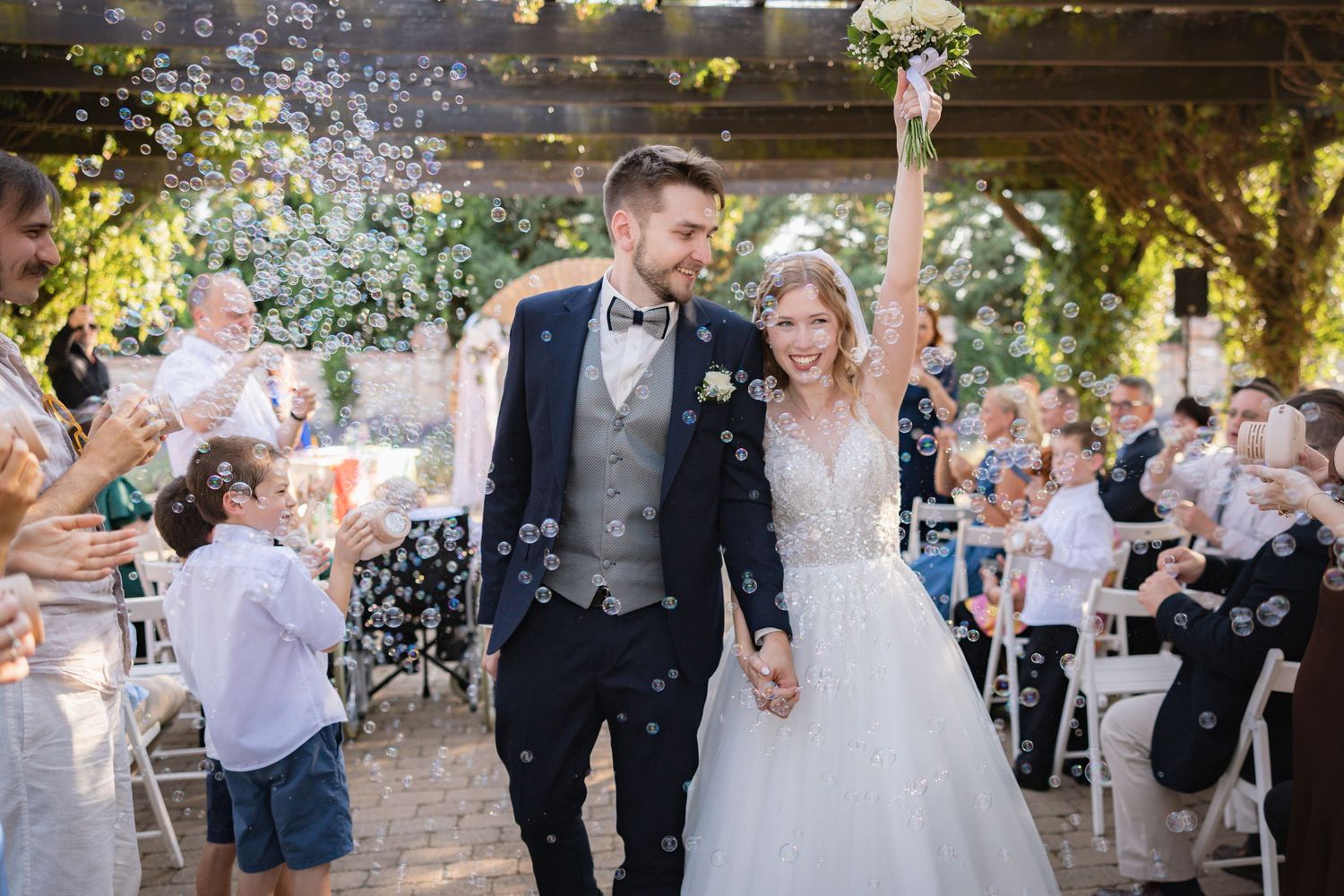 Bride and groom walk down aisle surrounded by bubbles and guests at an outdoor wedding ceremony.