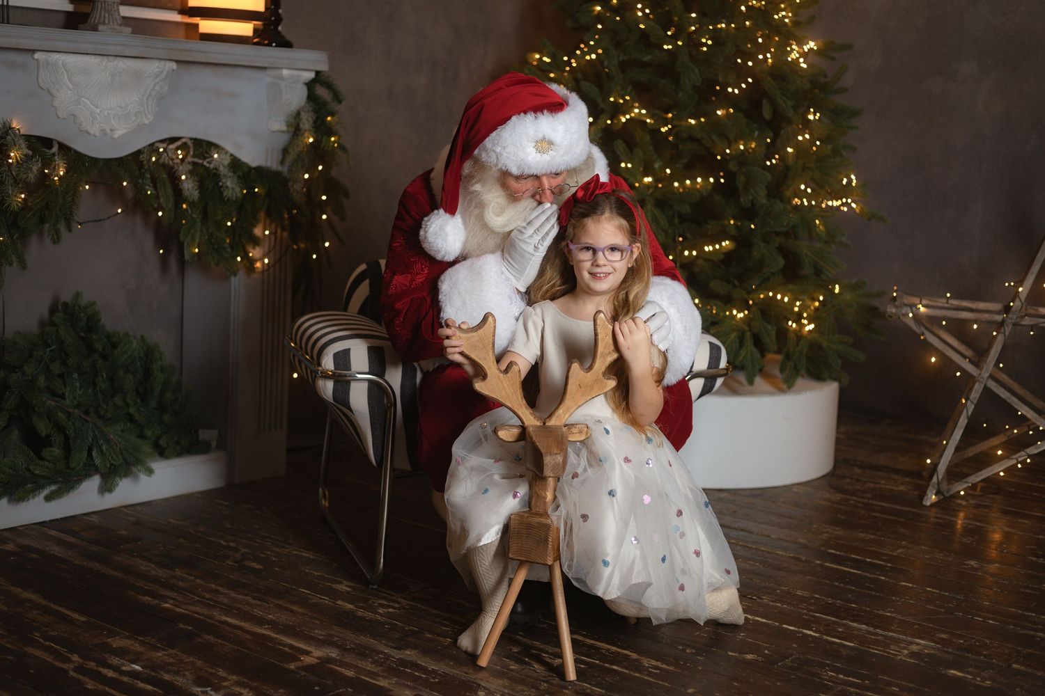 Santa Claus sits with a young girl beside a decorated Christmas tree, as she touches a wooden reindeer figurine.