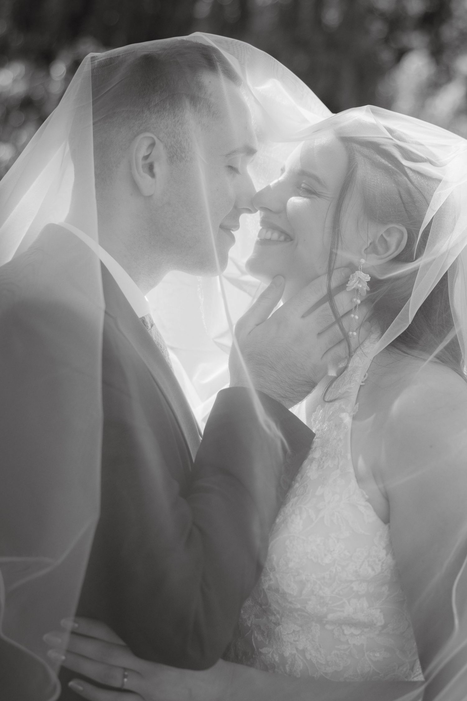 Black and white photo of smiling bride and groom sharing romantic moment under wedding veil.