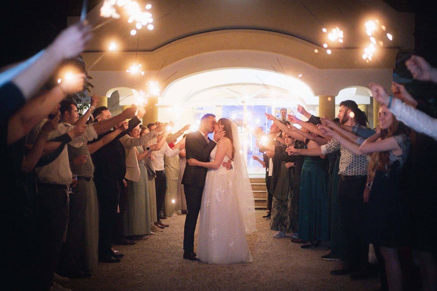 Bride and groom kissing under sparklers held by wedding guests during romantic nighttime sendoff ceremony.