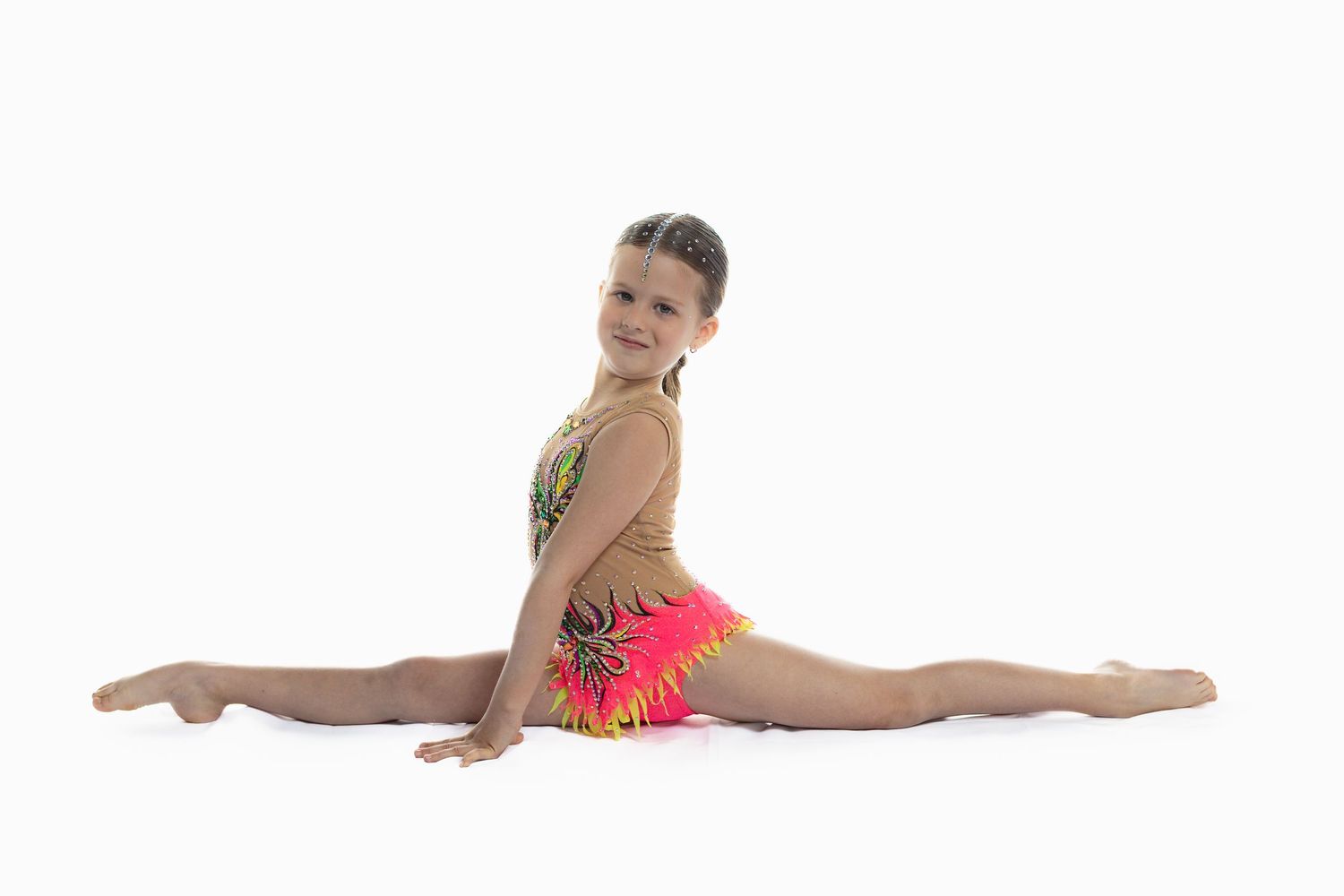 Young gymnast in colorful leotard performing splits on white background, smiling at camera.