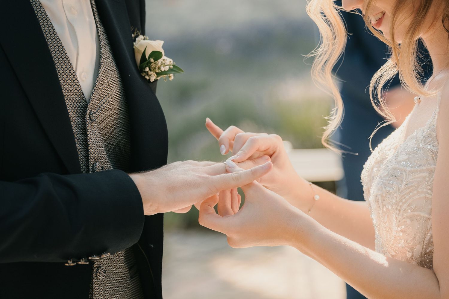Bride placing a wedding ring on groom's finger during ceremony, close-up of hands with boutonniere visible.