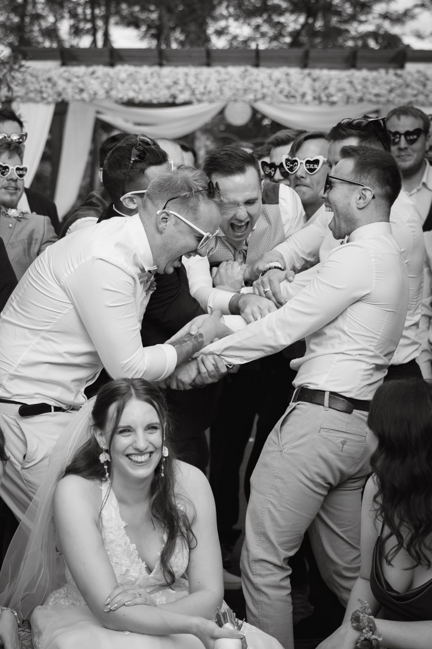 Black and white wedding photo of groomsmen celebrating joyfully while bride smiles in foreground at outdoor reception.