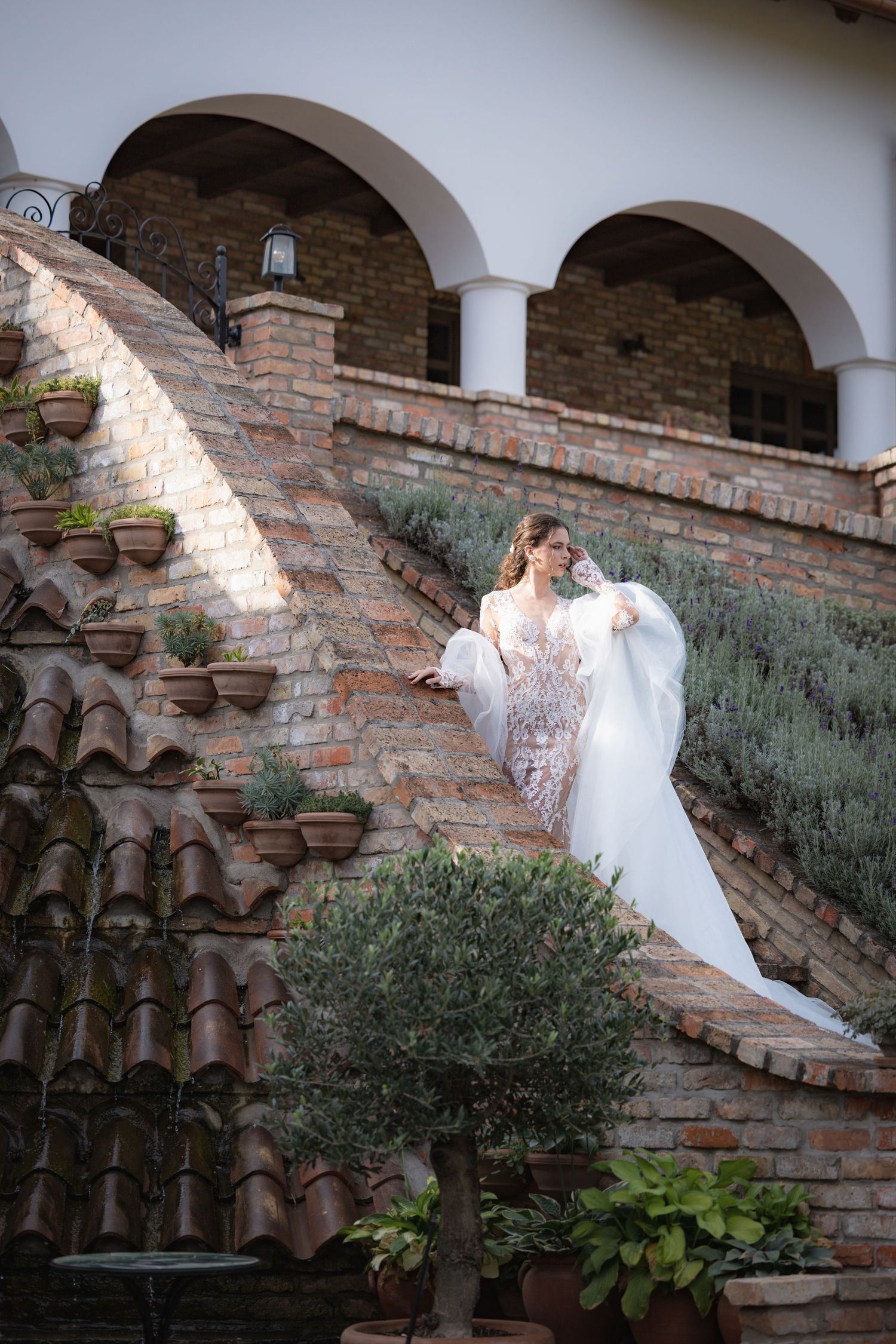 A person in a white dress descends rustic brick steps with arched columns and potted plants along the Mediterranean-style wall.